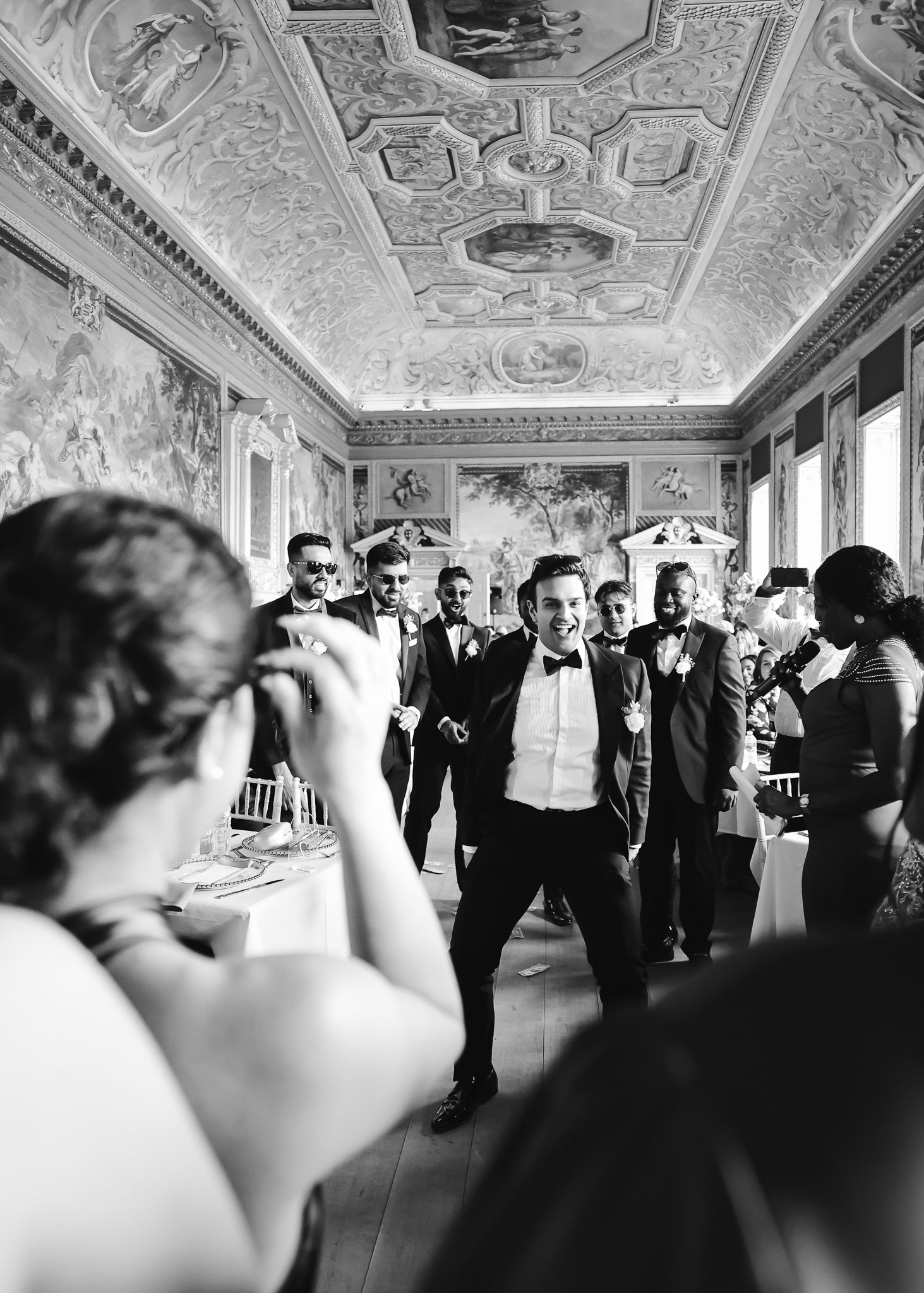 Guests in formal attire dance and socialize in an ornate ballroom with decorative ceiling frescoes and elegant architecture.