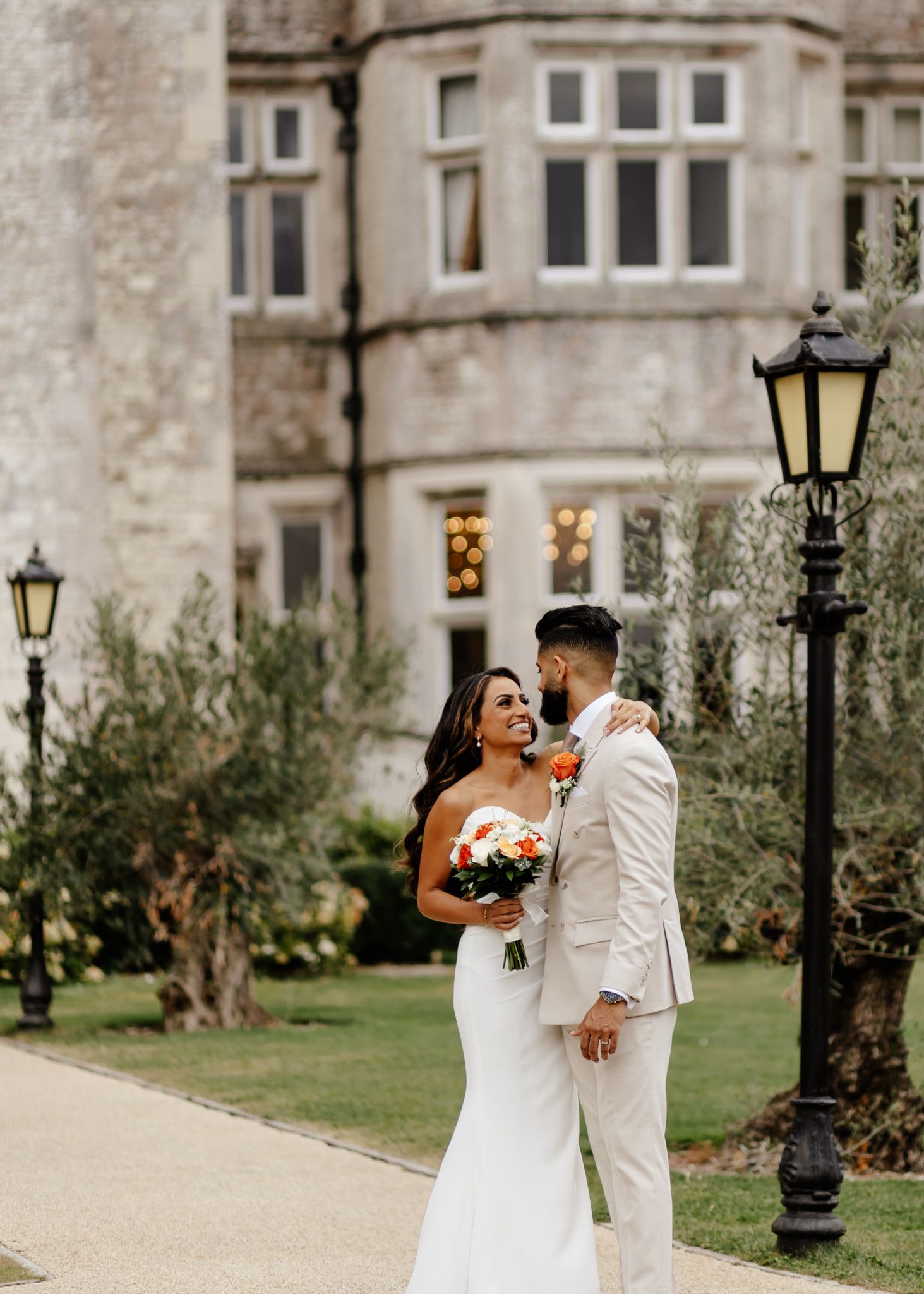 A couple in wedding attire shares a joyful moment together on a garden path lined with vintage lampposts and stone walls.