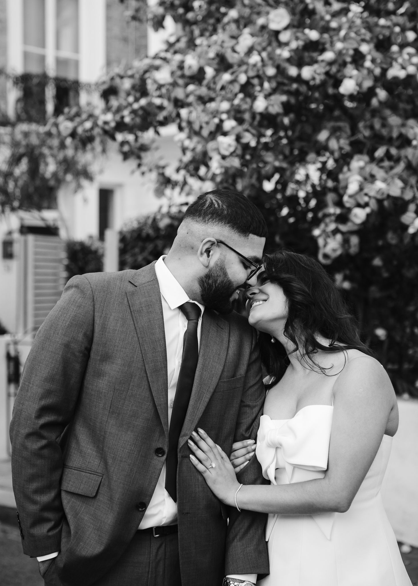 Black and white photo of a couple in wedding attire sharing a romantic moment outdoors near a flowering tree.
