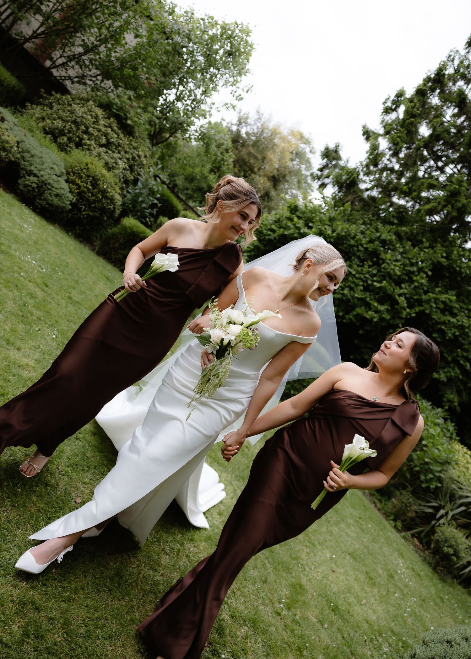 Bride in white gown walks with two bridesmaids in dark brown dresses, holding white bouquets in a lush green garden.