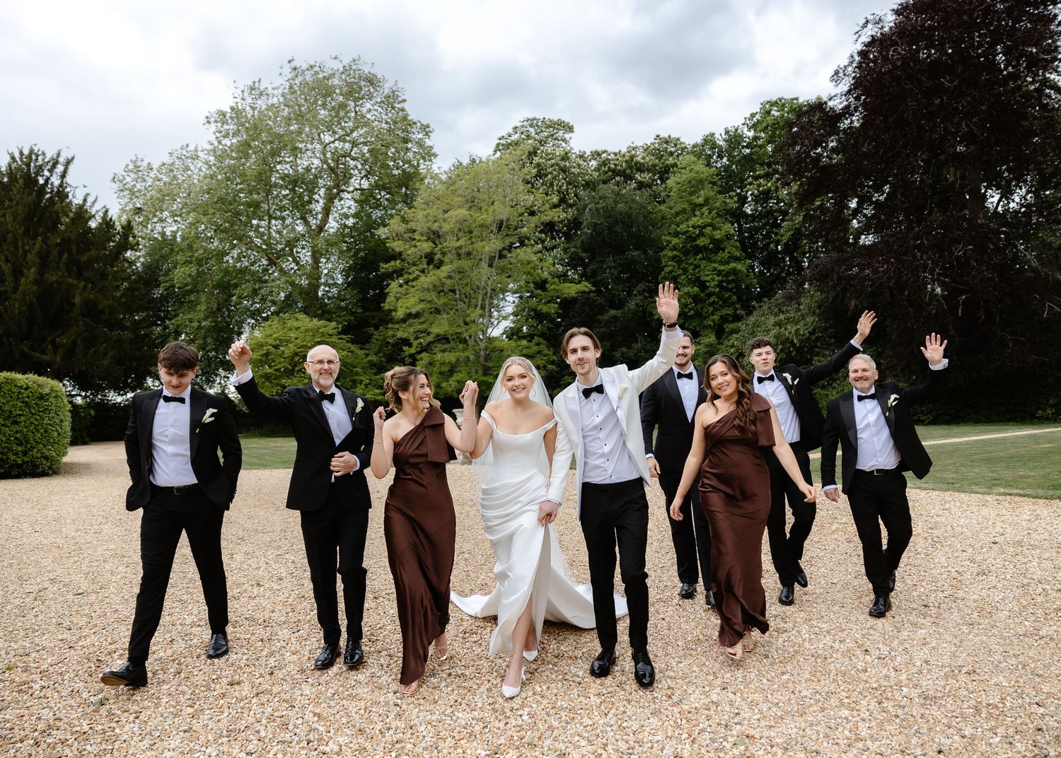 Wedding party celebrating outdoors, bride and groom leading groomsmen in tuxedos and bridesmaids in burgundy gowns.
