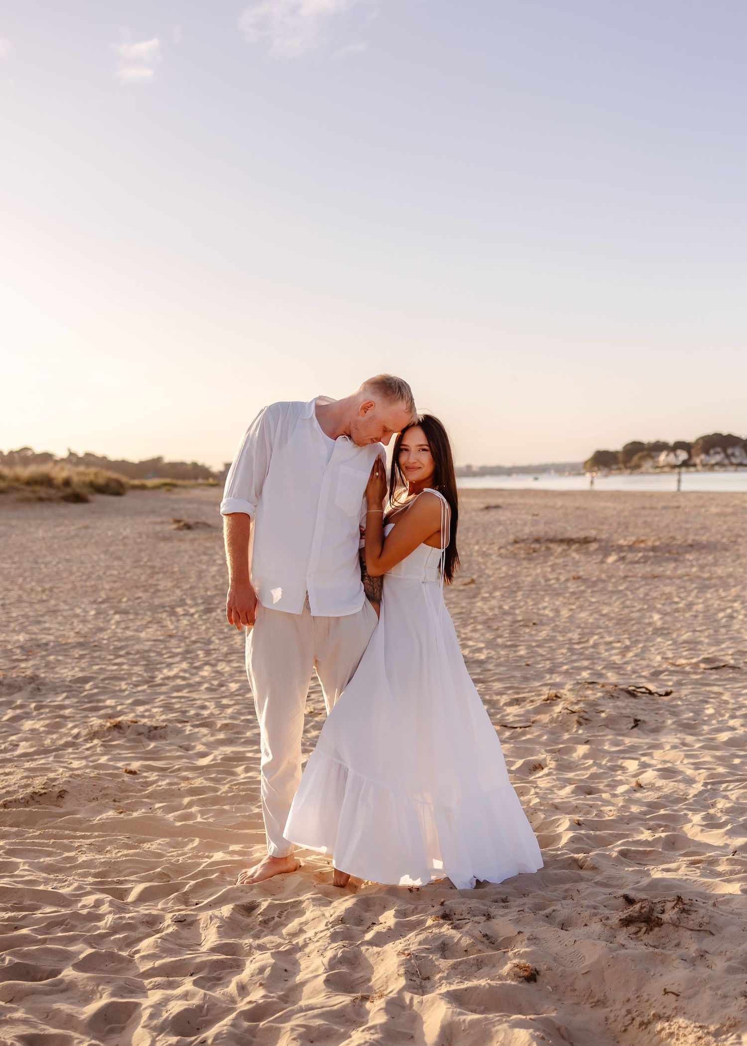 Couple in white outfits embracing on sandy beach at golden hour sunset.
