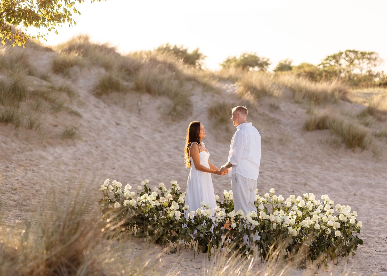 Couple in white attire holding hands on sandy beach dunes surrounded by white floral arrangement at golden hour.