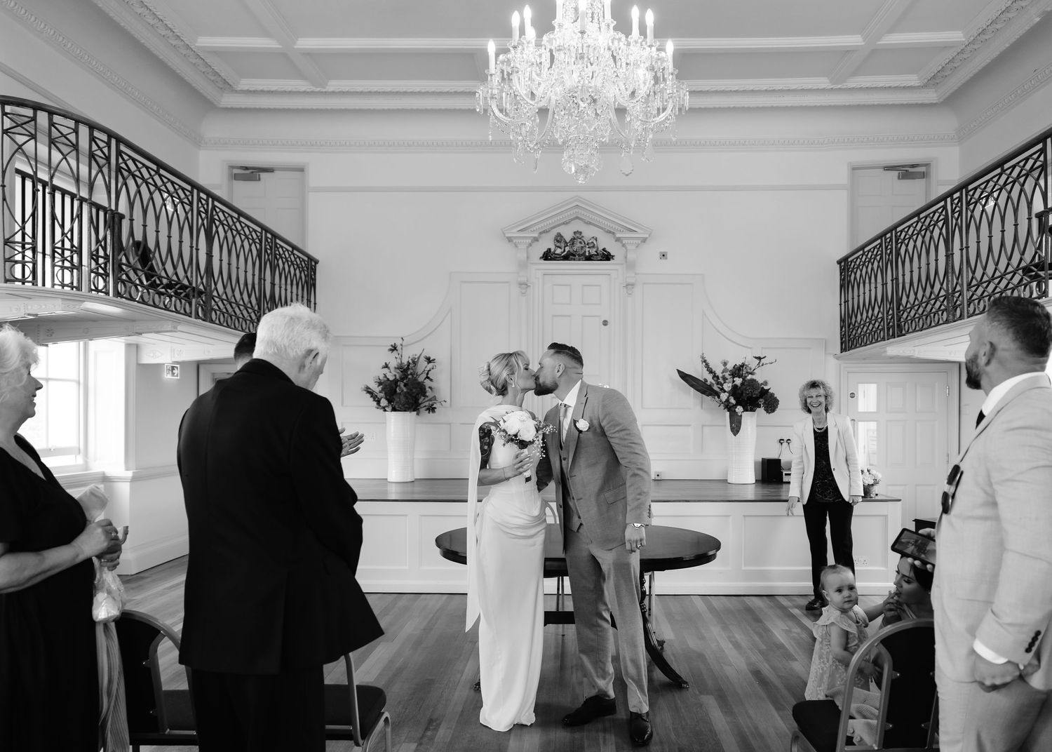 Newlyweds share first kiss under crystal chandelier in grand white wedding venue with balconies.