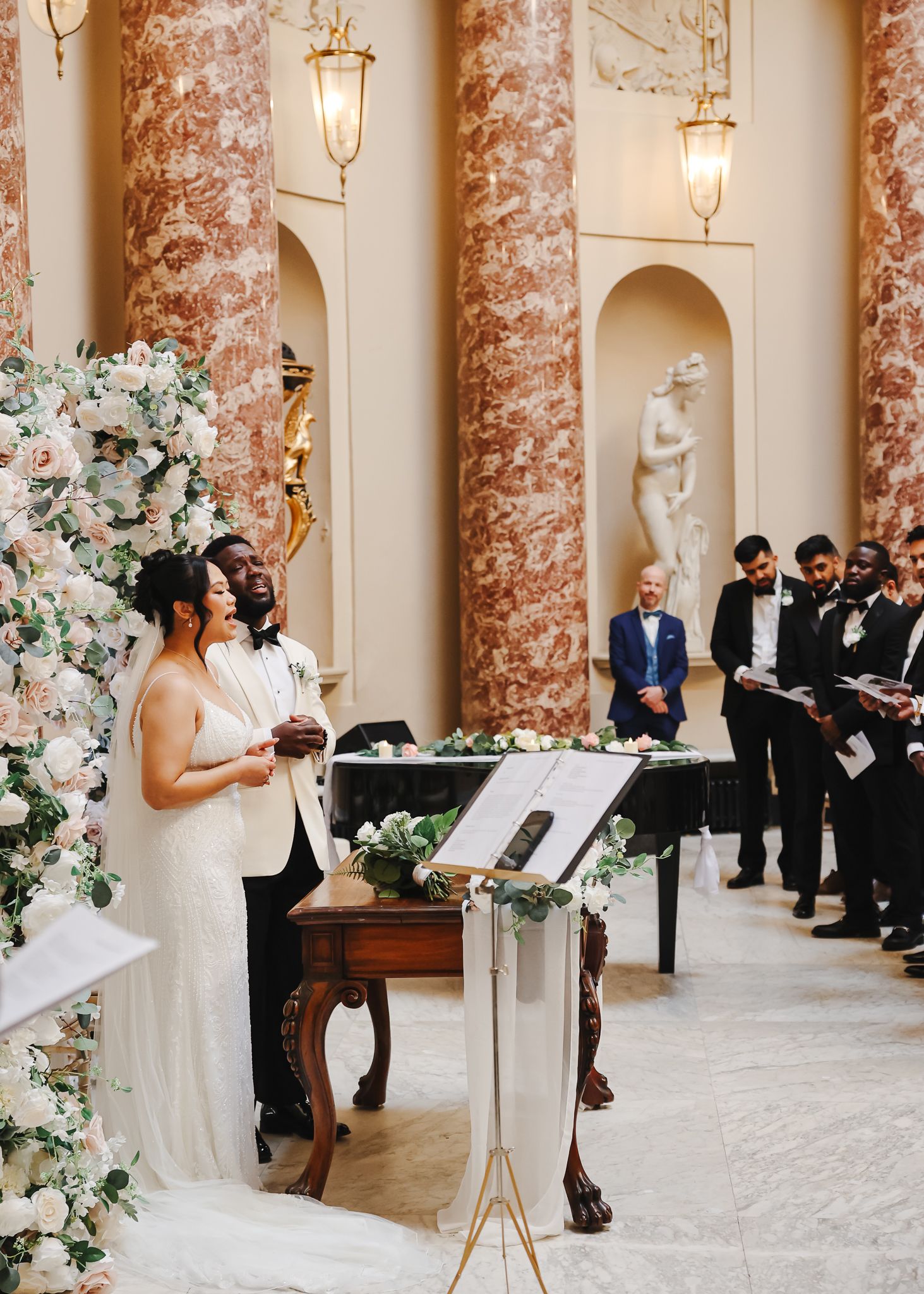 Elegant wedding ceremony in a marble columned venue decorated with white floral arrangements and hanging pendant lights.