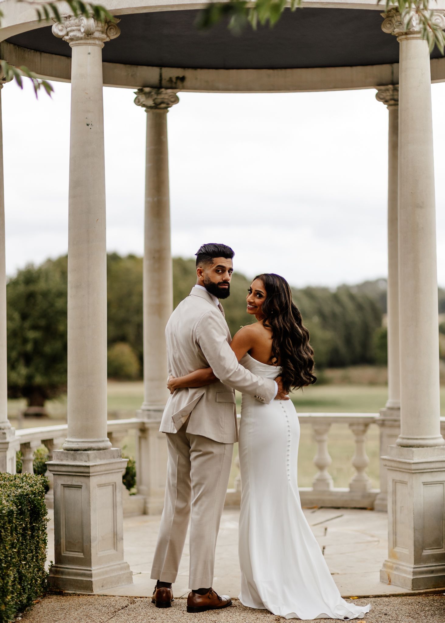 A wedding couple embraces under an elegant white gazebo with classical columns in an outdoor setting.