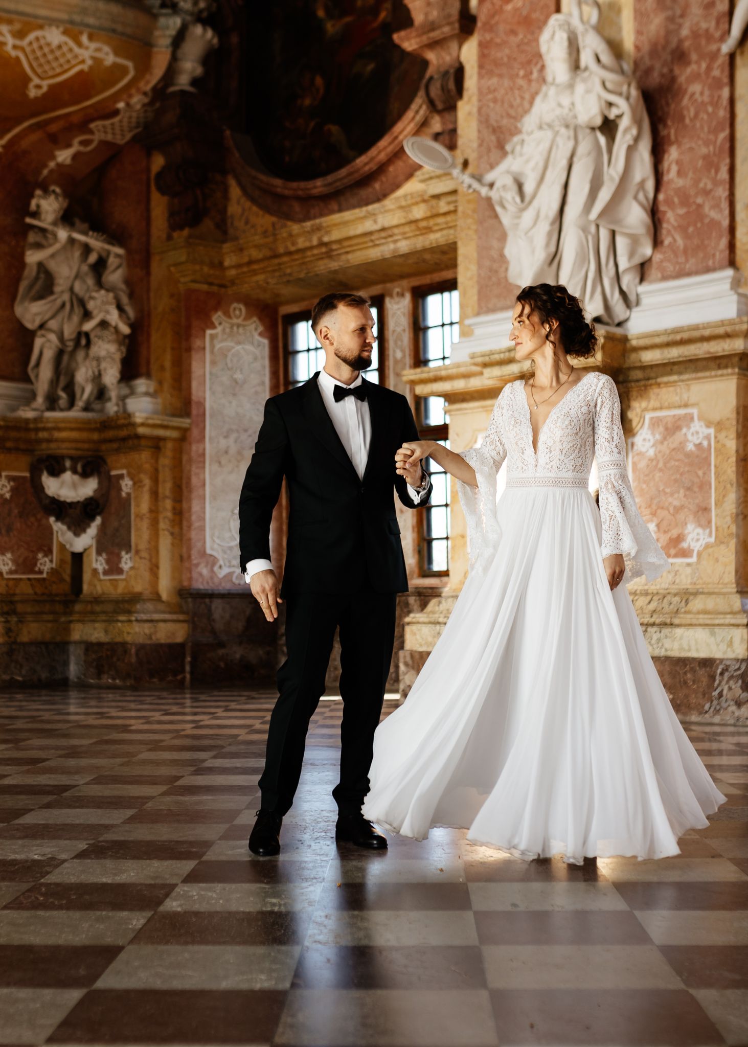 Elegant couple in wedding attire walking through a grand marble hallway with classical statues.