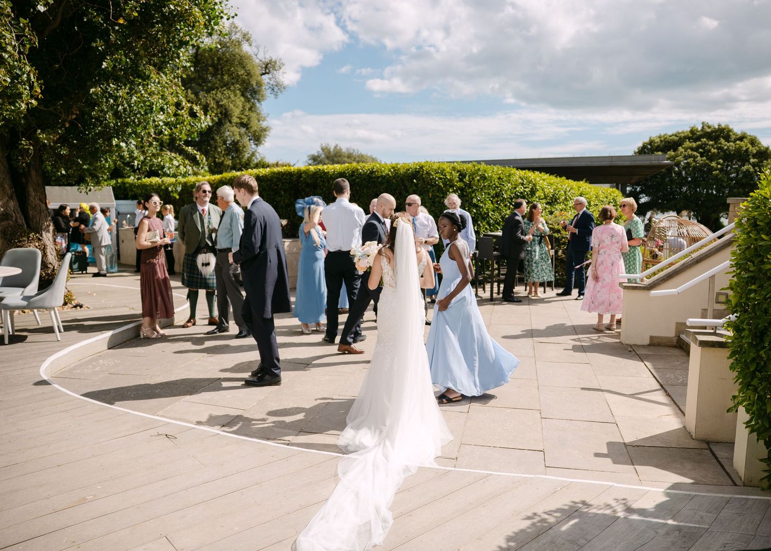 Bride in white gown with long veil mingles with wedding guests on sunny outdoor patio surrounded by lush hedges.