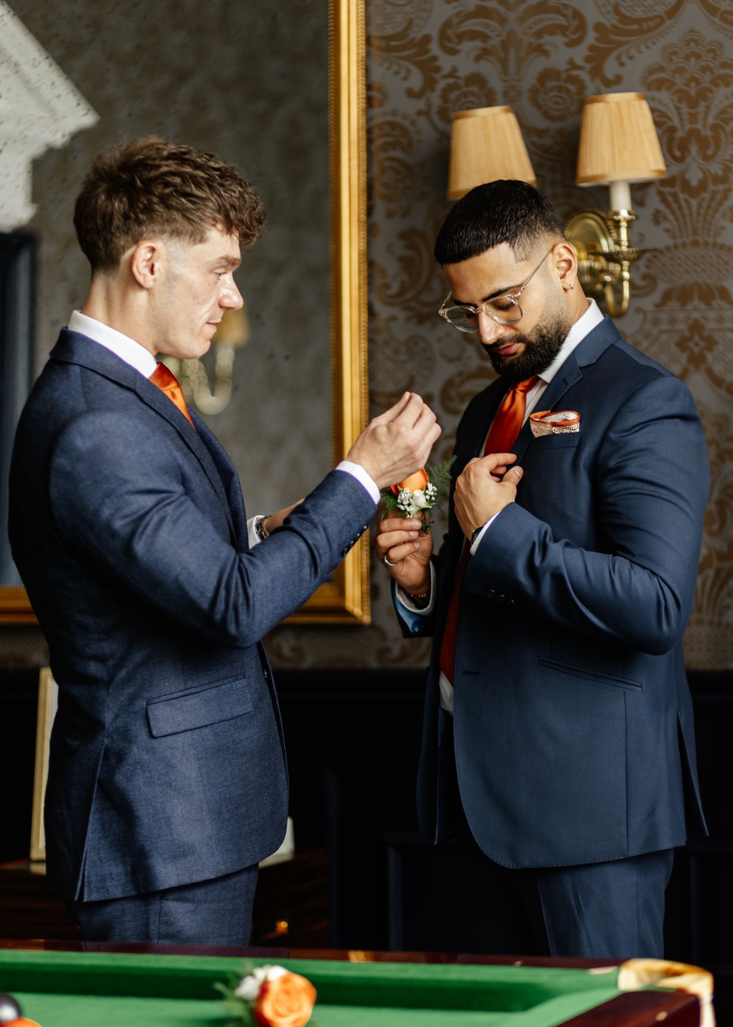 Two people in navy suits play snooker at an elegant venue with ornate wall decorations and sconces.