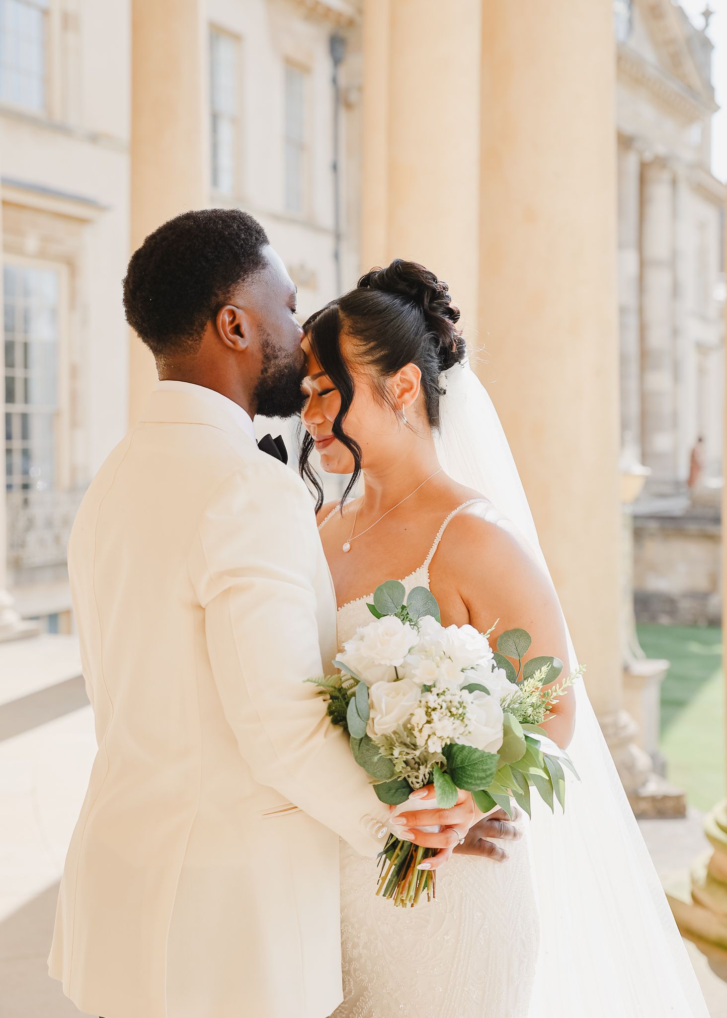 A wedding couple shares an intimate moment on a grand columned porch with elegant white and peach floral bouquets.