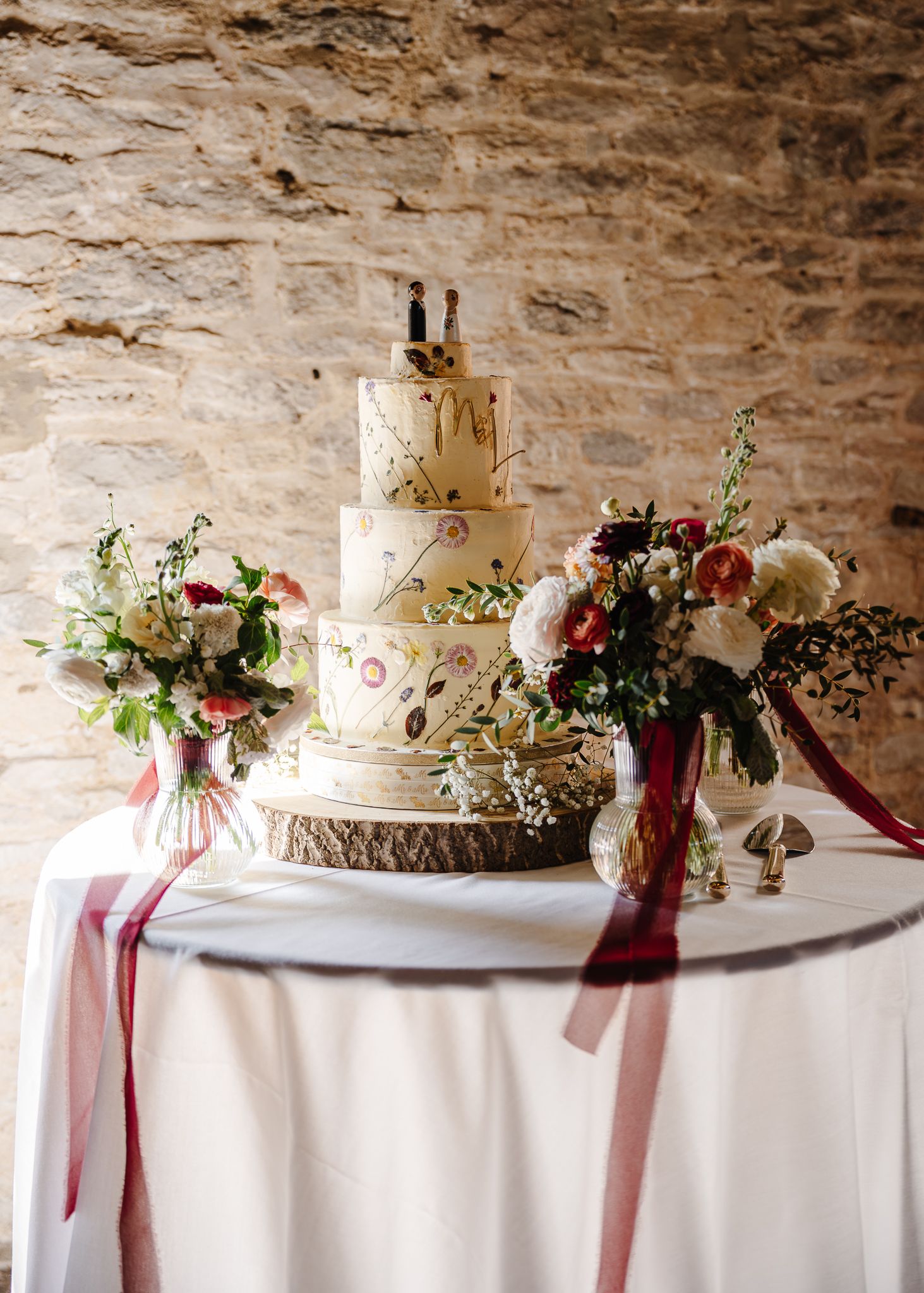 Elegant white wedding cake with fresh flowers and burgundy ribbons displayed on rustic stone table against stone wall.