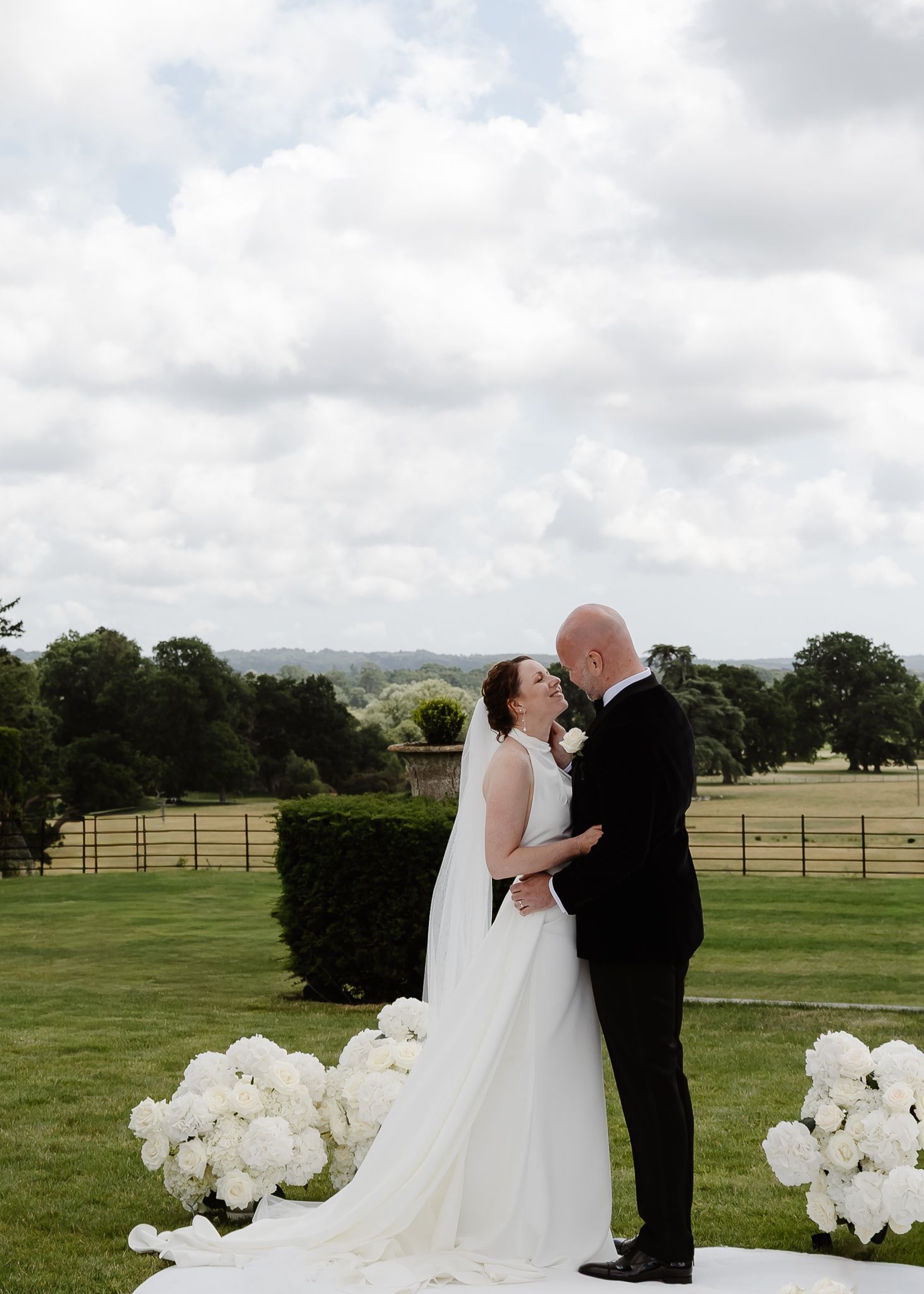 A romantic wedding couple shares a tender kiss on a scenic hilltop with rolling green fields and white floral decor.