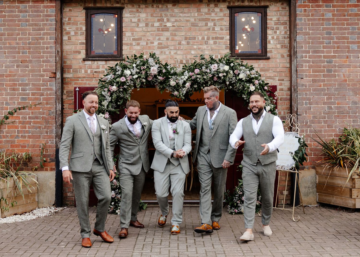 Groomsmen in gray and beige suits pose casually on brick patio during wedding photoshoot.
