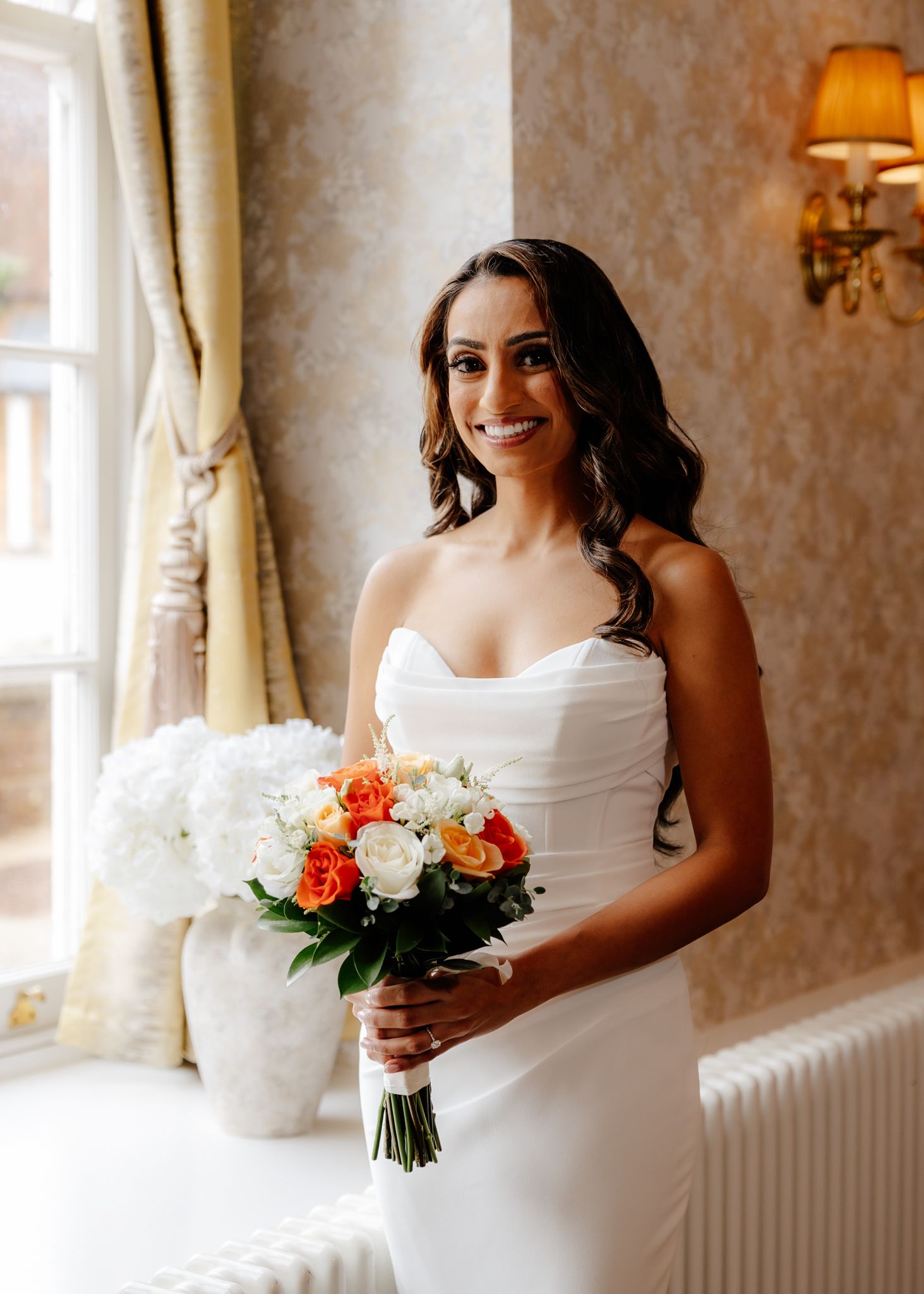 A bride in a strapless white wedding dress holds a bouquet of coral and white flowers by a sunlit window.