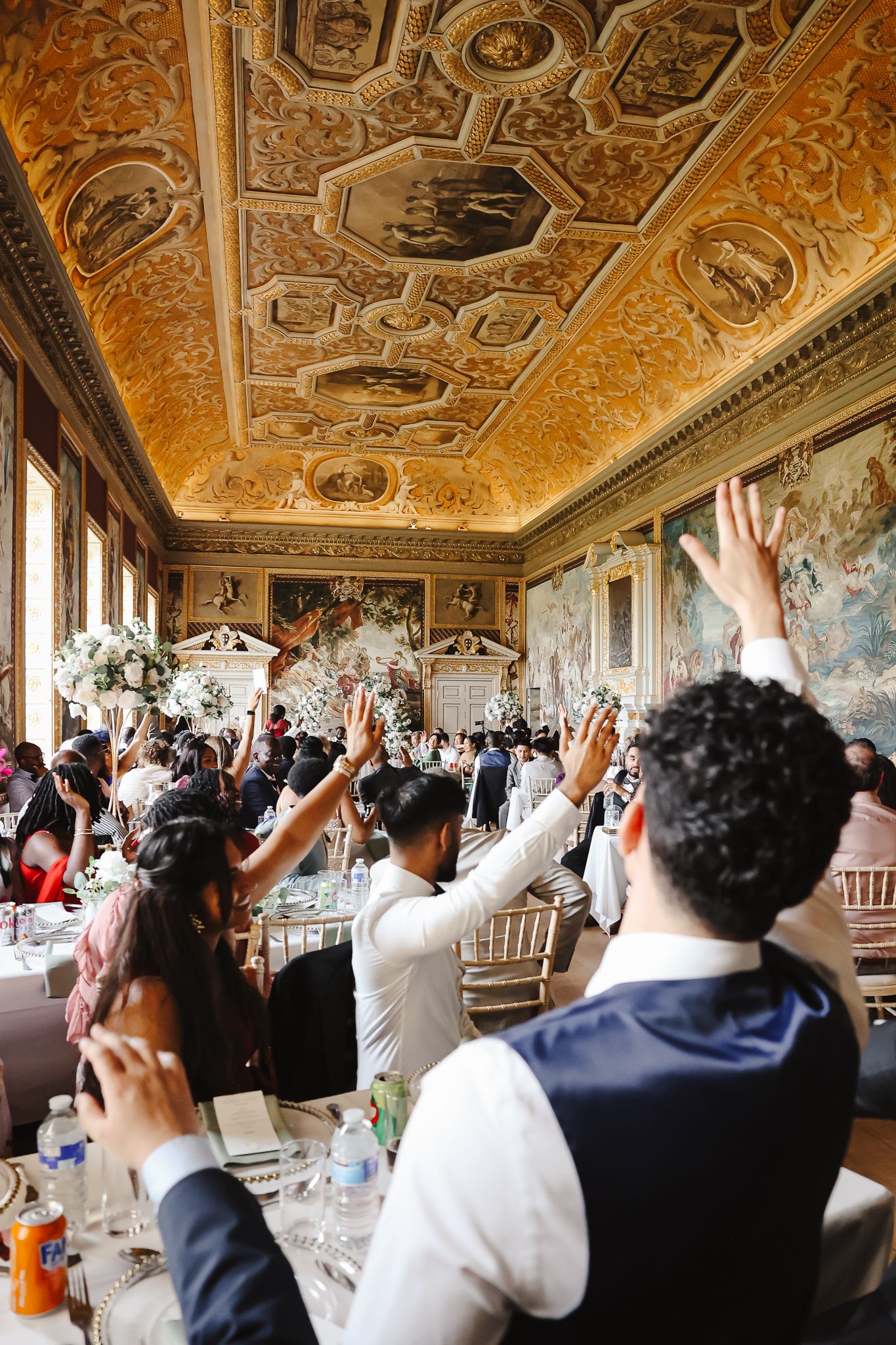 Wedding guests raise their hands in celebration under an ornately painted gold ceiling at a reception venue.
