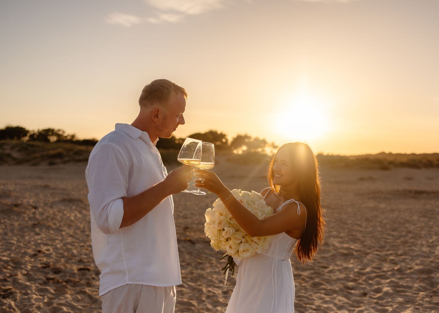 Couple toasting wine glasses on a sandy beach at golden sunset, woman holding white flowers in romantic celebration.