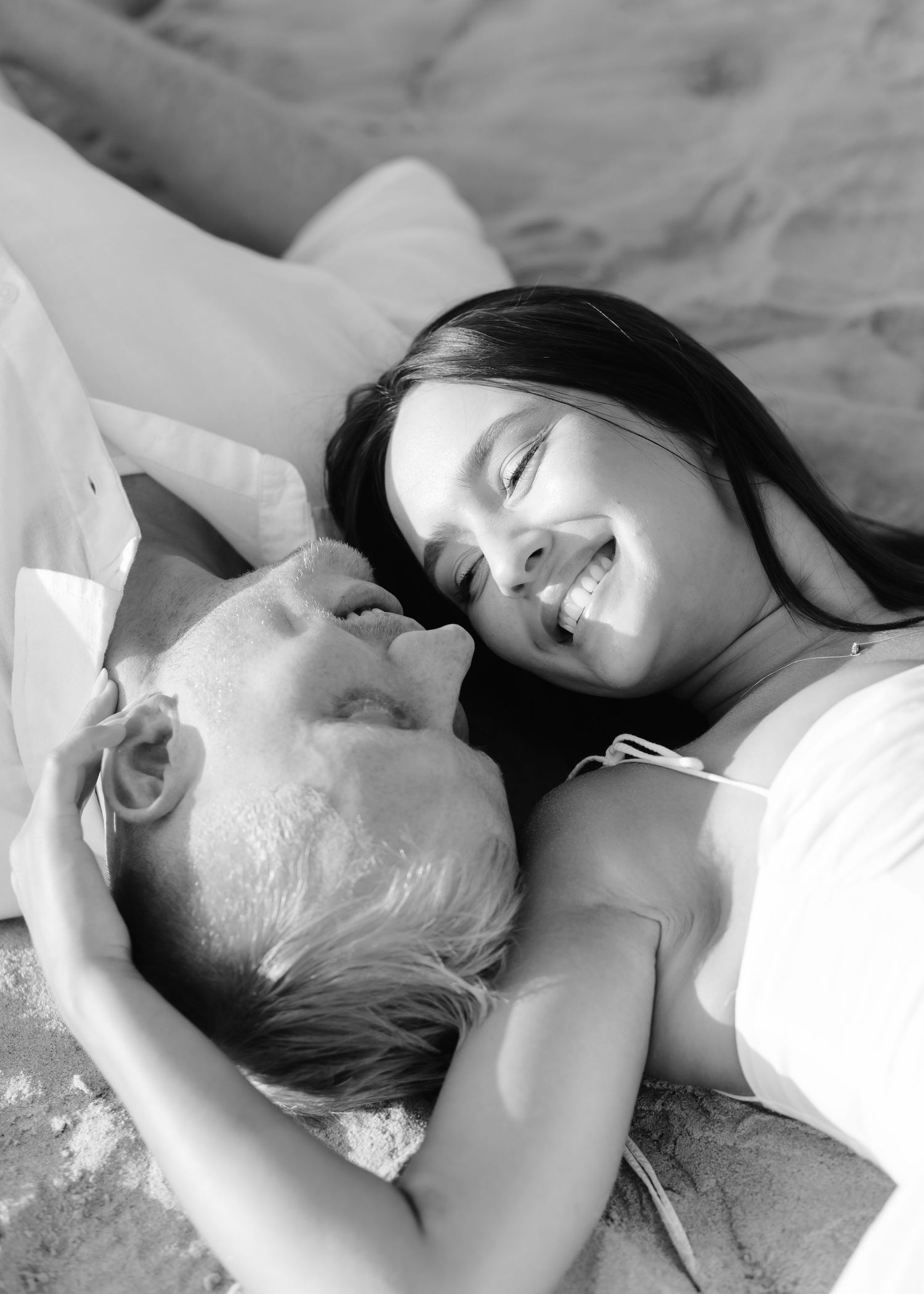 Black and white photo of smiling couple lying together on sandy beach.