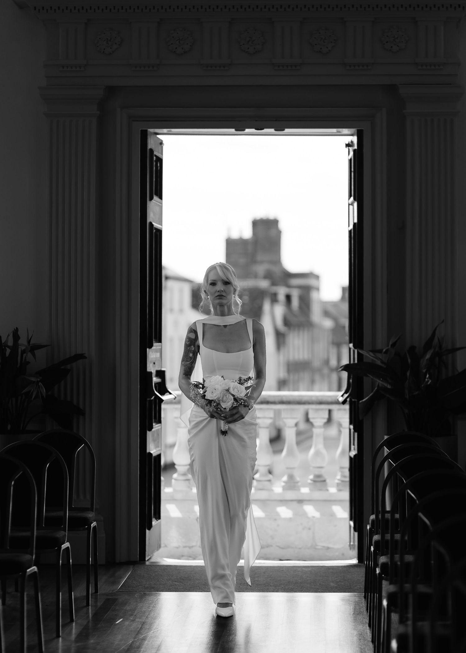 Black and white photo of a bride in a sleek white gown walking through elegant double doors with a bouquet.