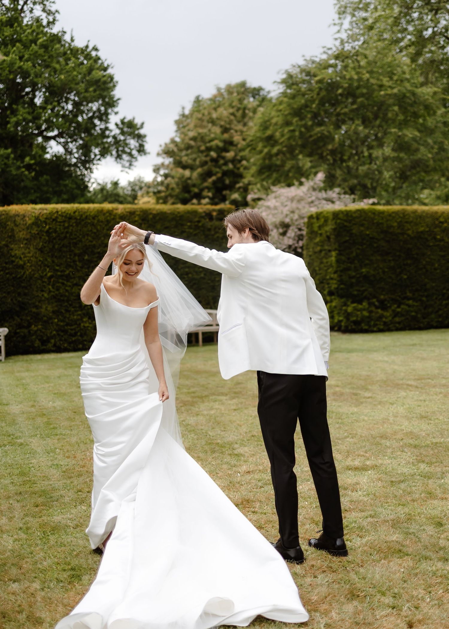 Groom twirls bride in white mermaid gown and flowing veil on lush green lawn at outdoor wedding celebration.