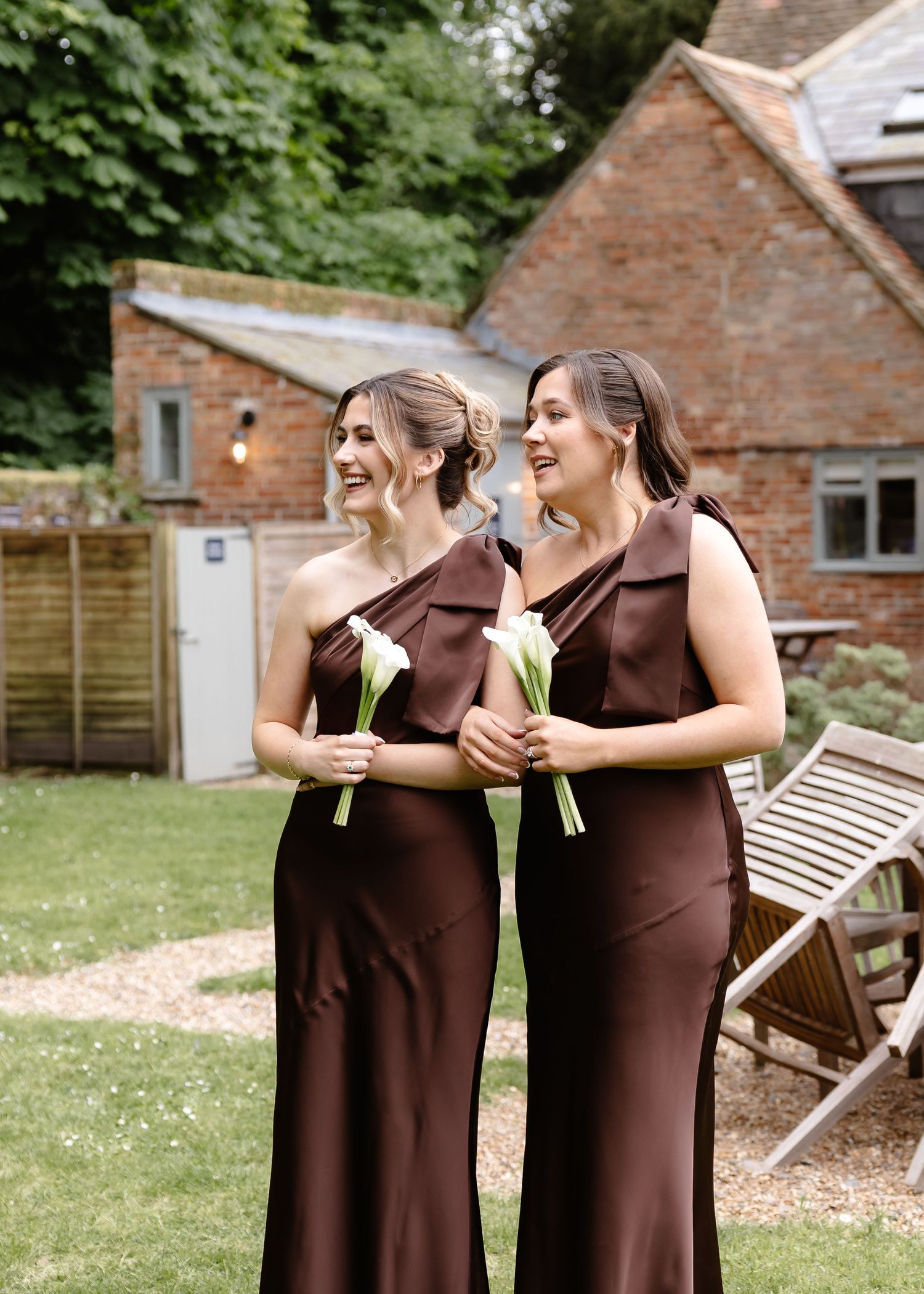 Two bridesmaids in elegant brown satin one-shoulder gowns holding white calla lily bouquets outdoors at a wedding venue.