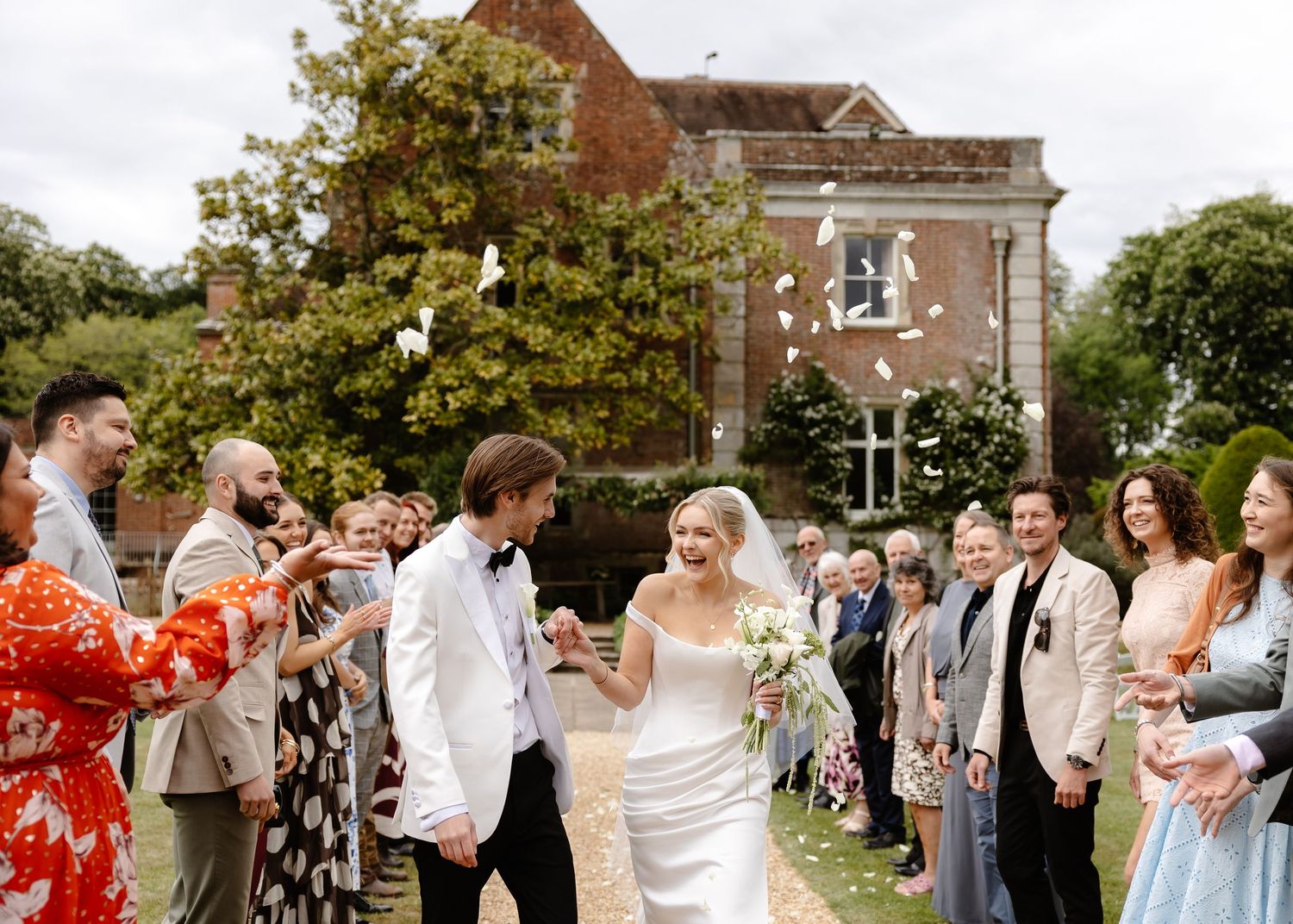 Bride and groom walk through cheering guests throwing confetti outside an elegant English country house wedding venue.