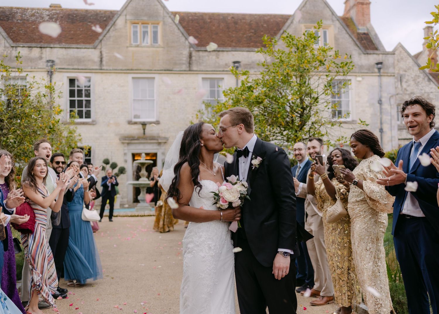 Newlywed couple sharing a kiss outdoors at a wedding venue while guests celebrate around them.
