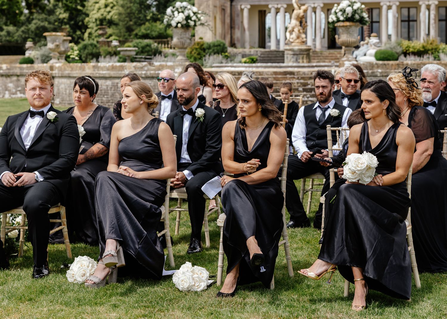 Wedding party members in black attire sit together on chairs arranged outdoors on a lawn.