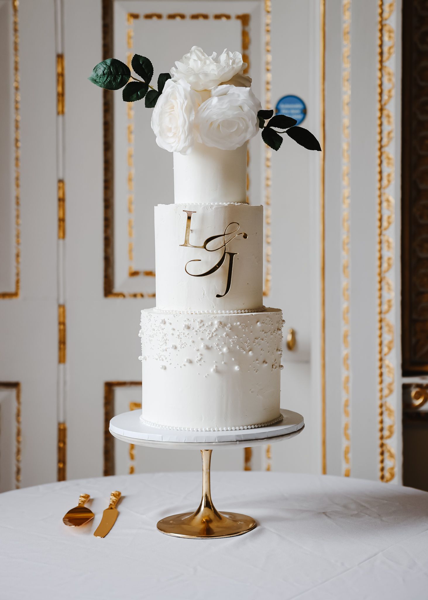 Elegant white wedding cake with delicate floral decorations and gold accents displayed in an ornate room.