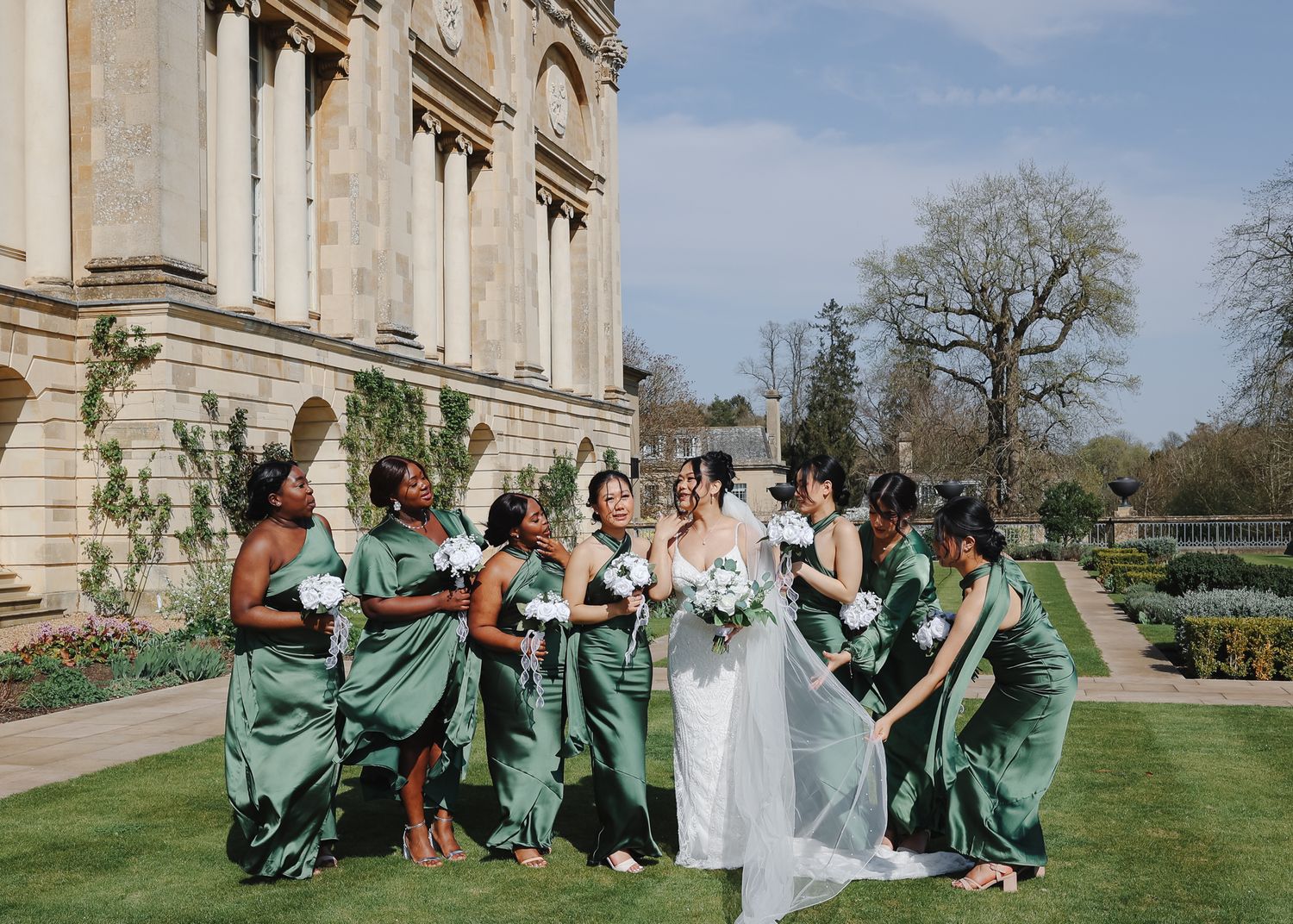 Wedding party members in mint green dresses sit on grass outside a grand stone building on a sunny day.
