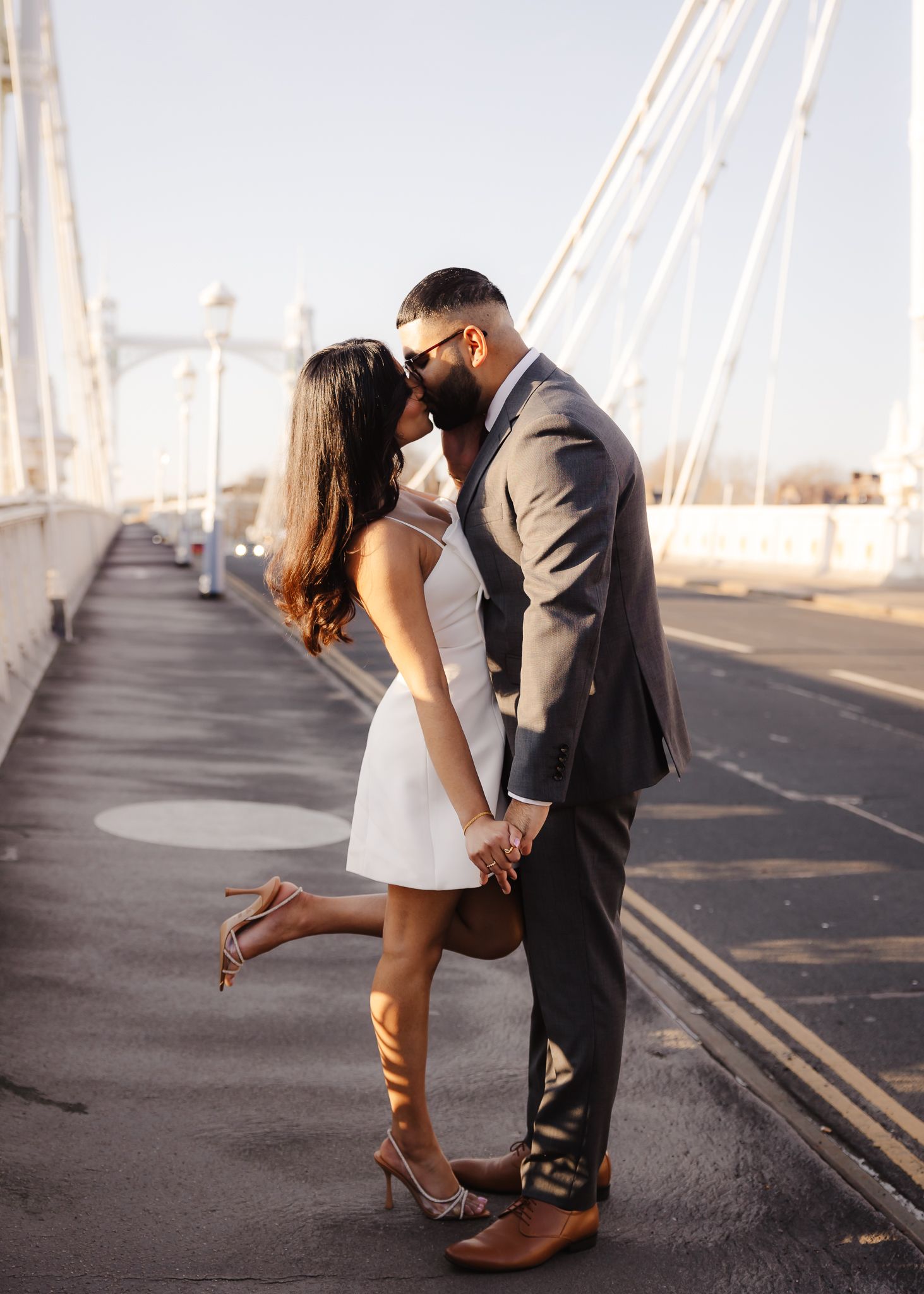 Couple sharing a kiss on a bridge at golden hour, woman in white mini dress and heels.