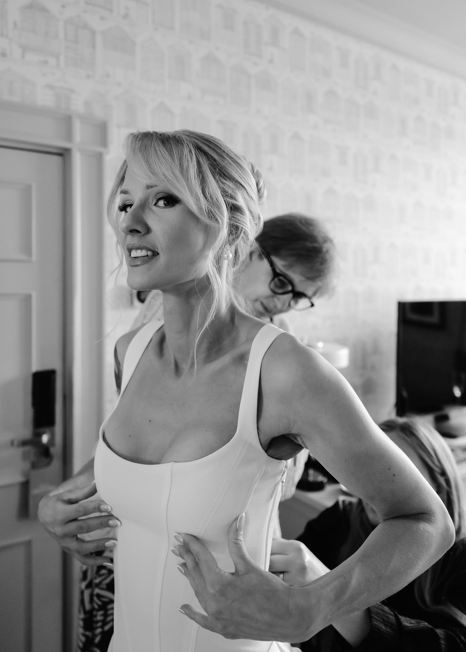 Black and white photo of a blonde bride in a white dress being helped by a woman during wedding preparations.