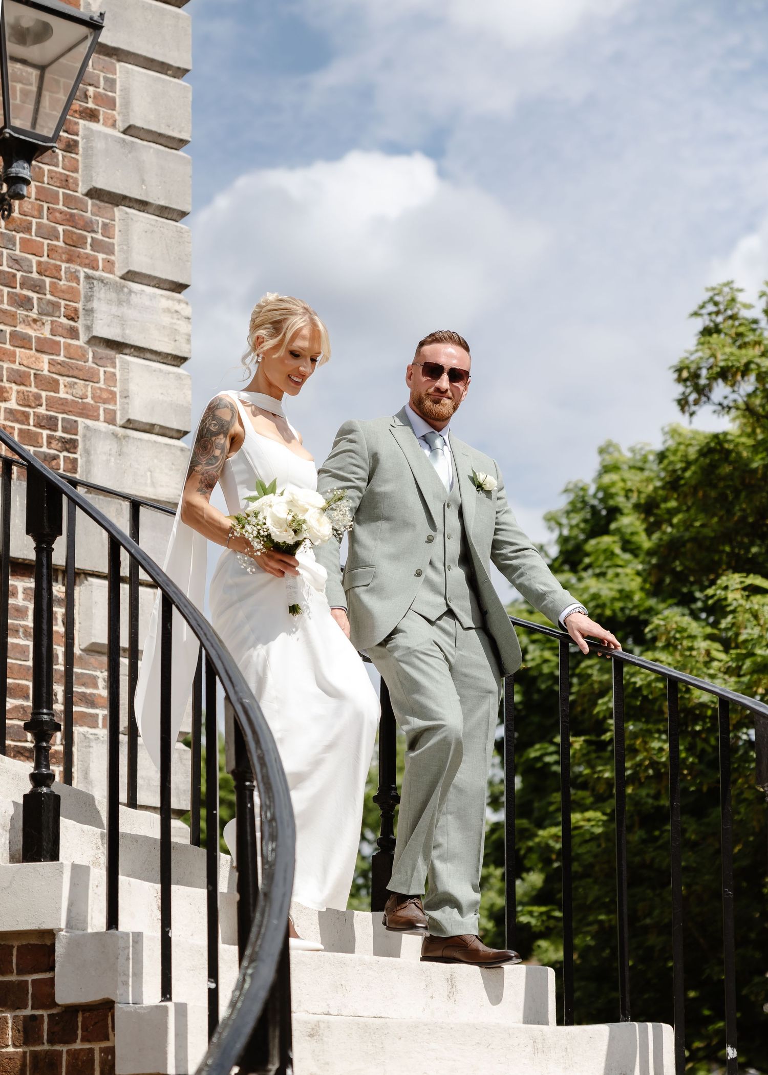 Bride and groom descend stone steps at a brick venue, she in a white gown with bouquet, he in a sage suit.