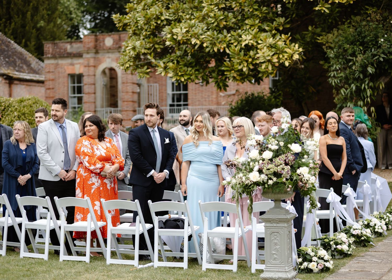 Guests standing at an outdoor wedding ceremony with white chairs and floral arrangements in a garden setting.
