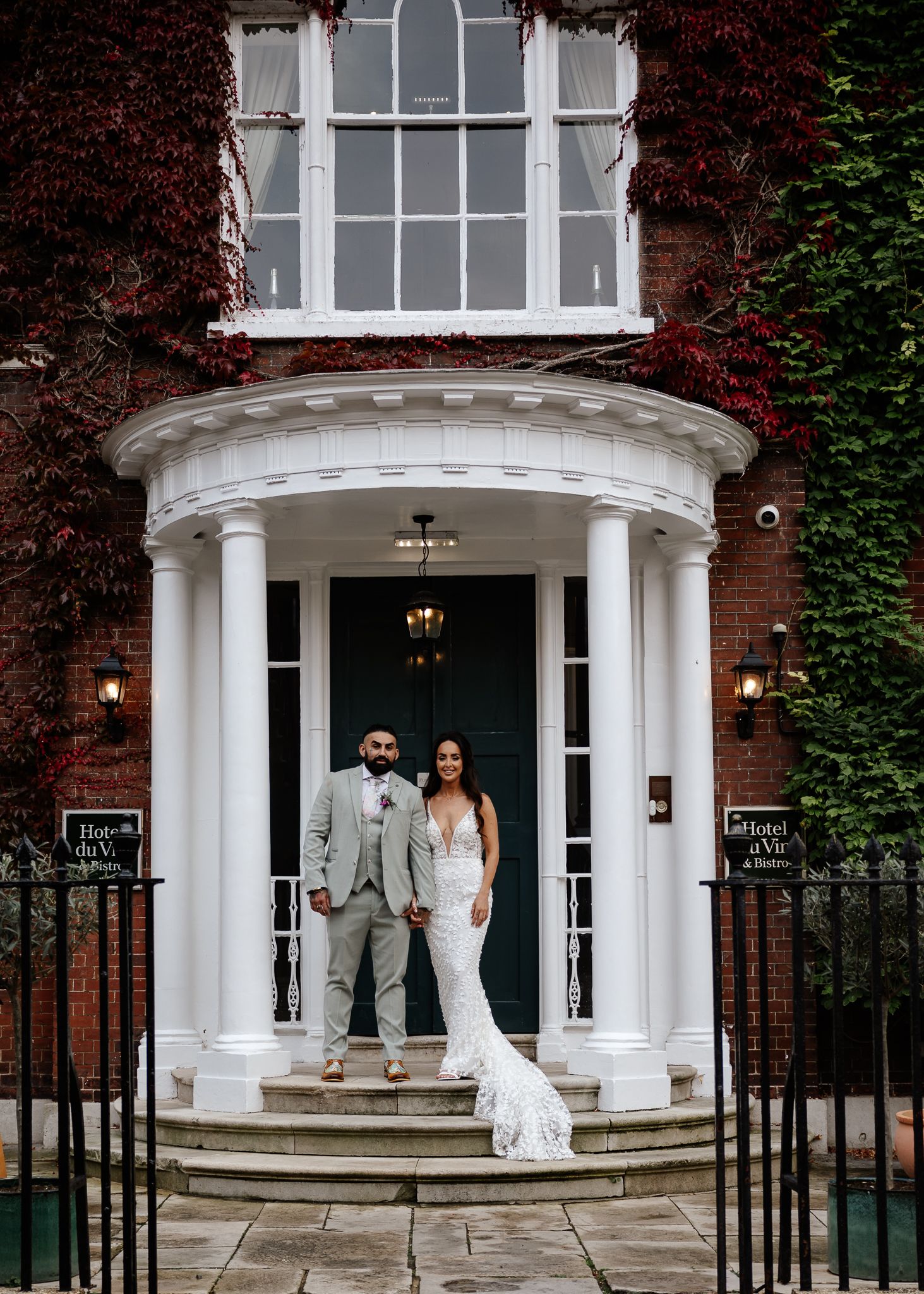 A wedding couple stands in front of an elegant Georgian entrance with white columns and red brick walls.