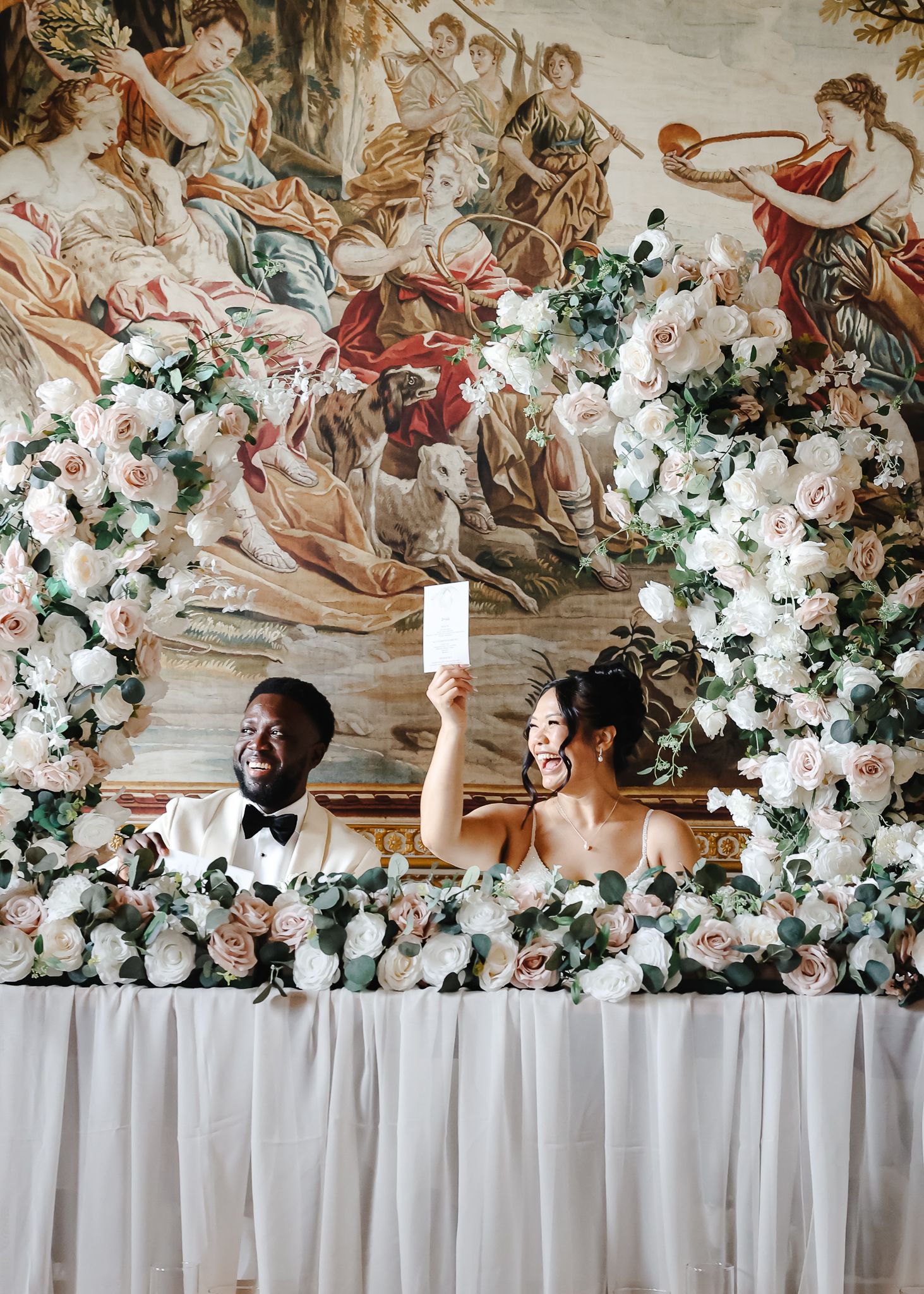 Wedding party stands at elegant head table decorated with lush white floral arrangements and ornate wall mural backdrop.
