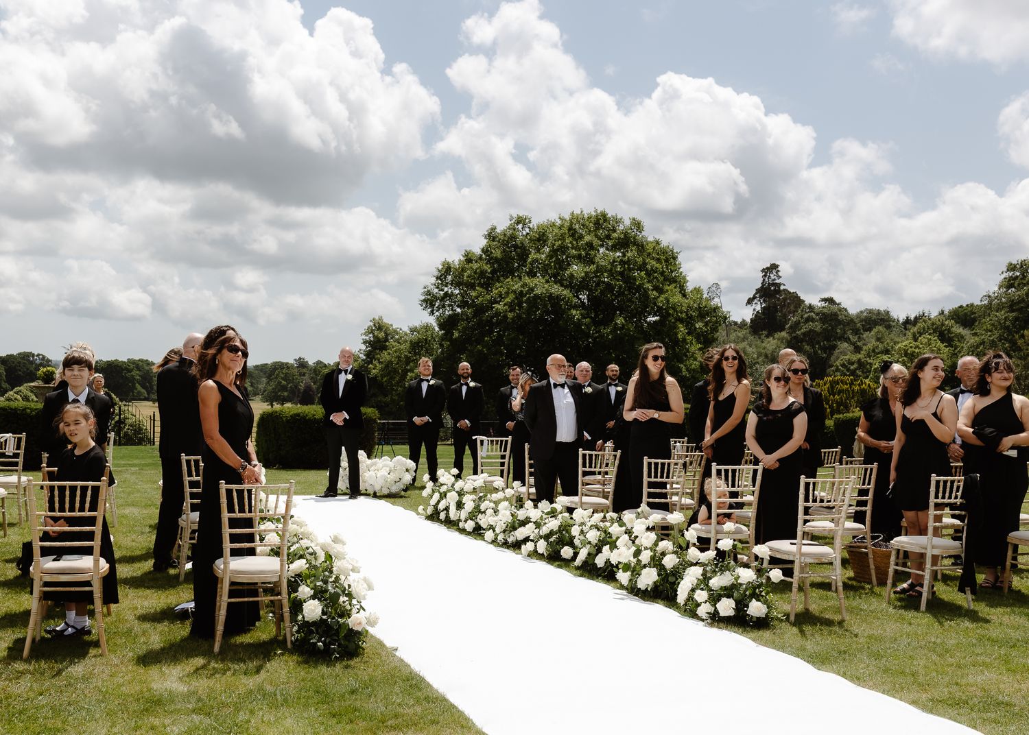 Wedding ceremony setup on a sunny lawn with white floral aisle decorations and guests in black attire.