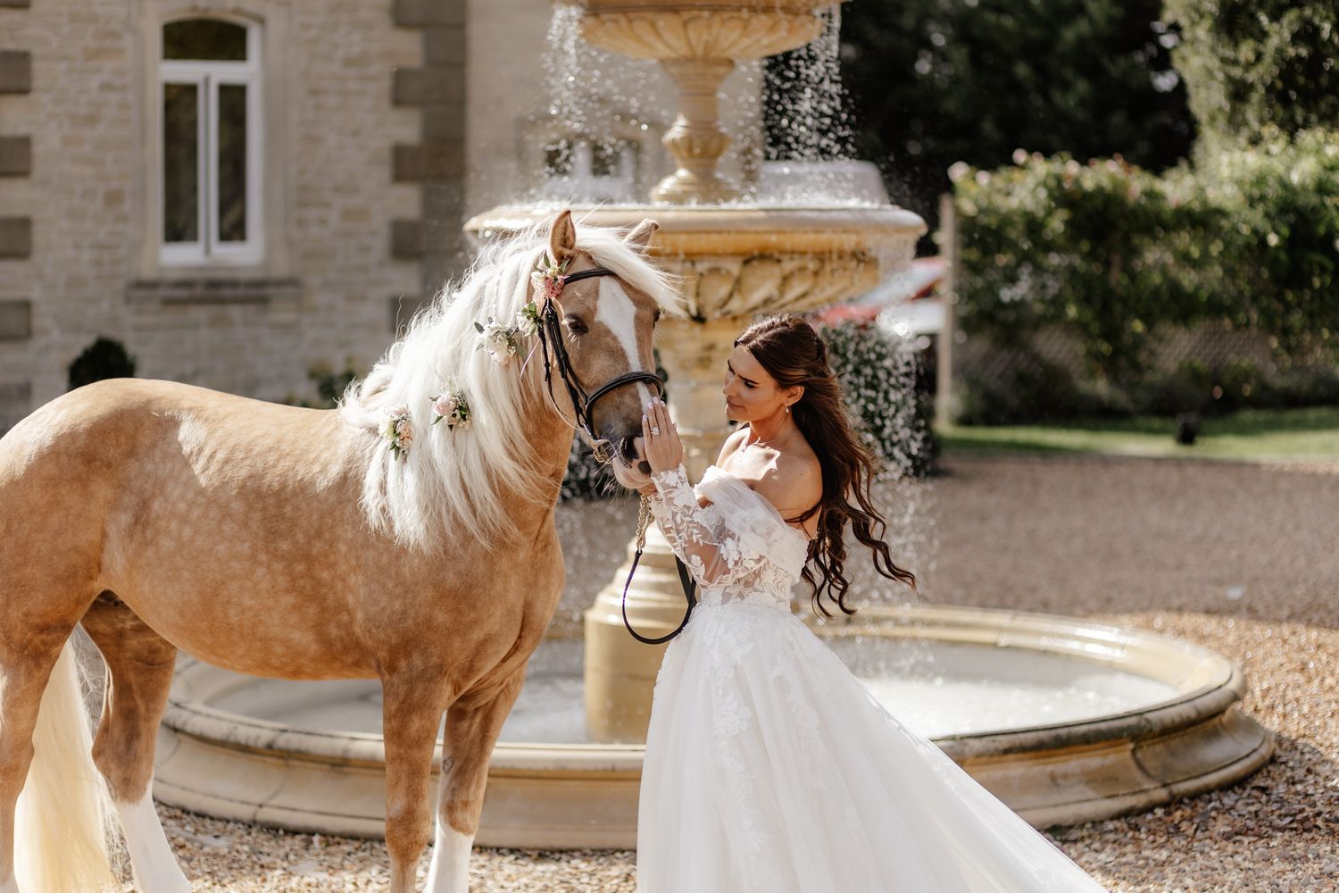 A palomino horse and person in a white dress stand together in front of a stone building during a wedding photoshoot.