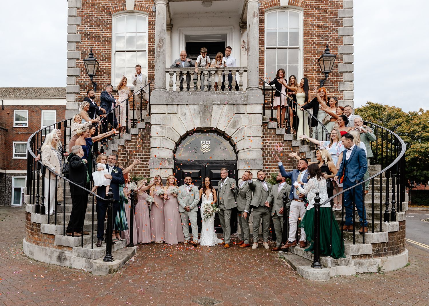 Wedding party gathers for formal group photos on brick steps outside historic building entrance.
