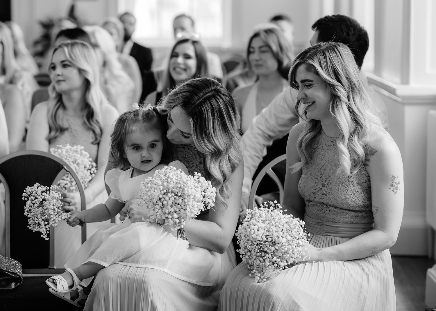Black and white photo of bridesmaids in elegant dresses holding baby's breath bouquets during a wedding ceremony.