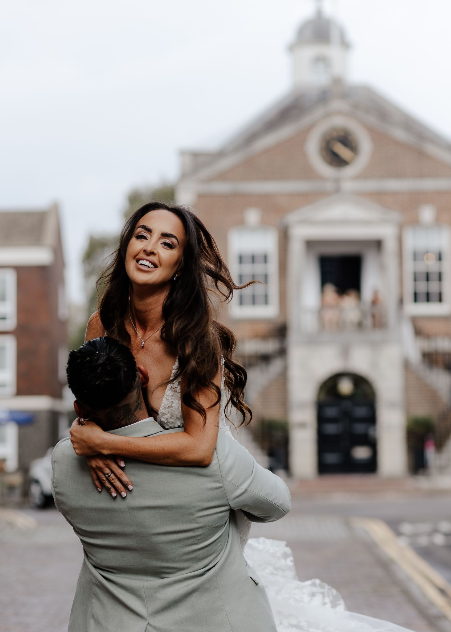 A couple embraces and laughs joyfully outside a traditional brick building on their wedding day.