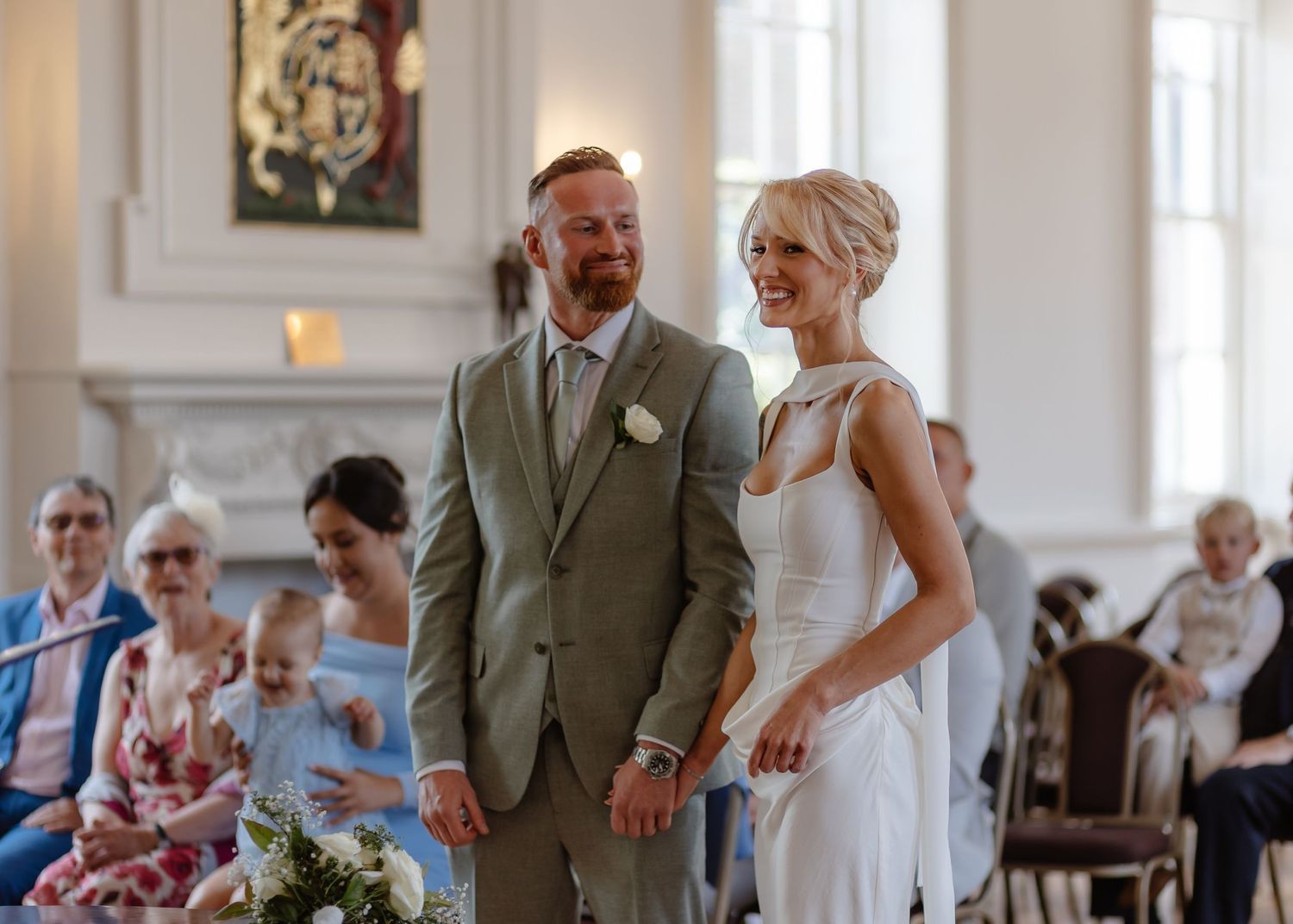 Smiling bride and groom stand together during elegant wedding ceremony with seated guests in bright white venue.