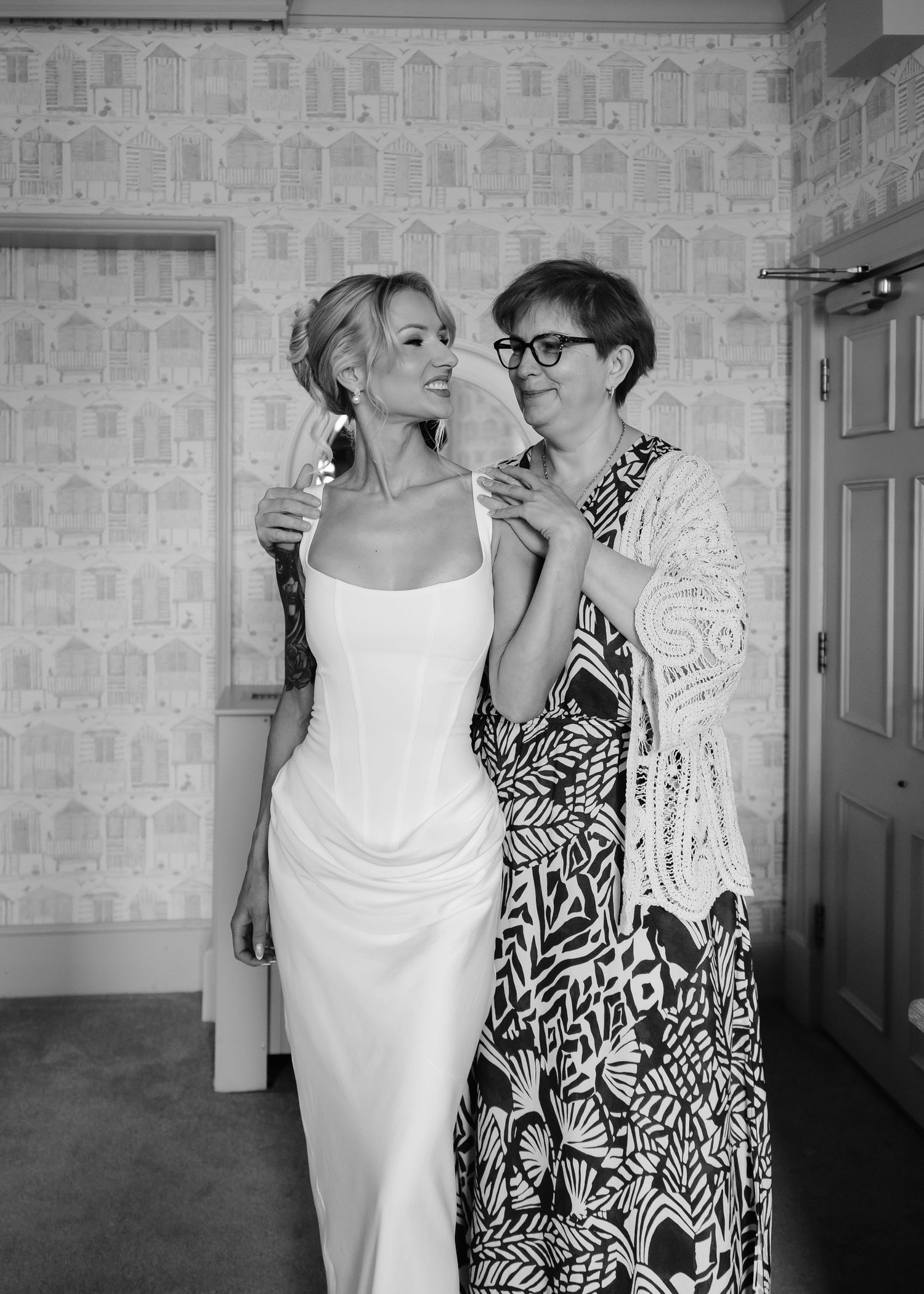 Black and white photo of a bride in a white gown smiling with an older woman in a patterned dress.