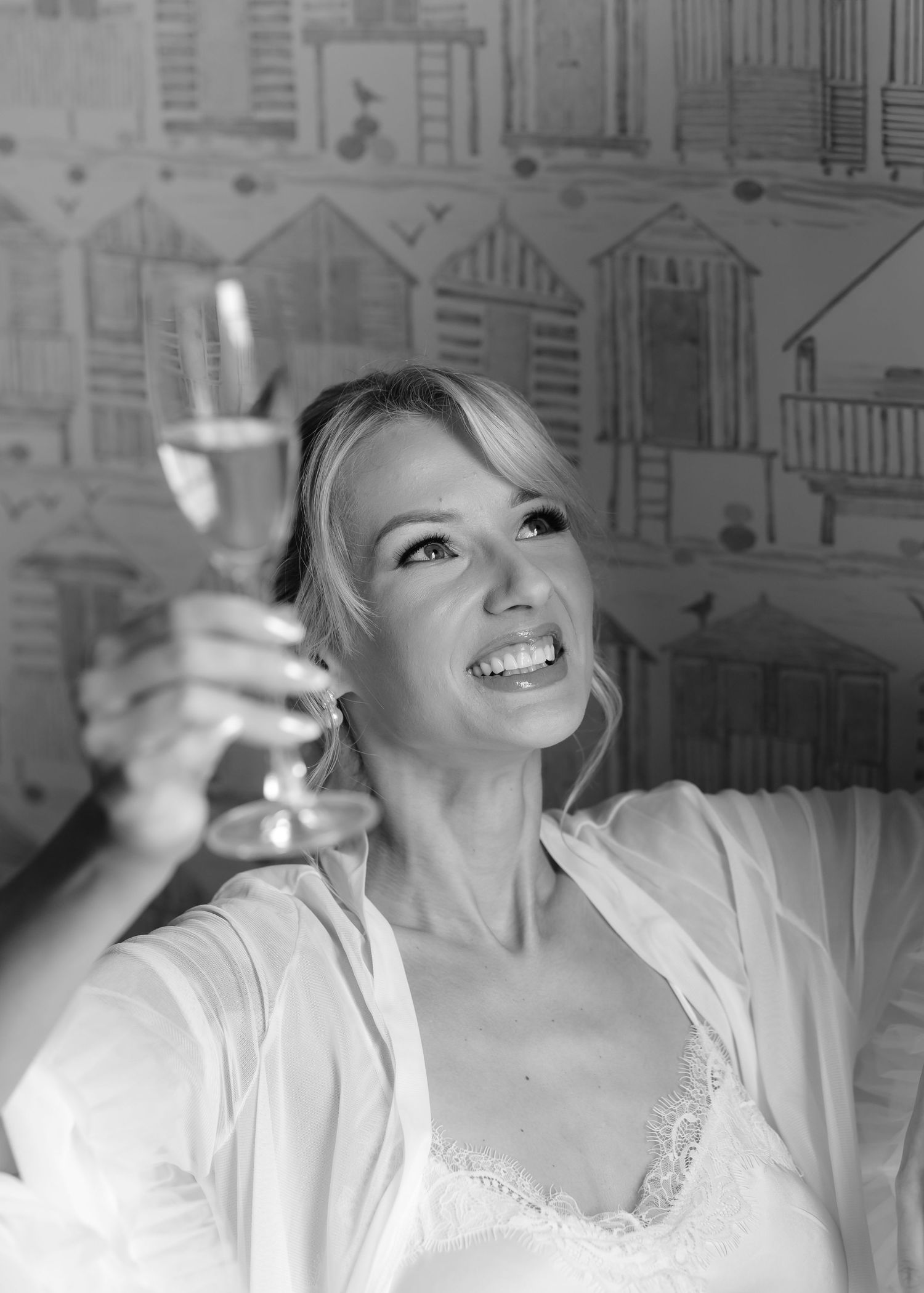 Smiling blonde woman in white robe raising a champagne glass, black and white photo with beach hut wallpaper.