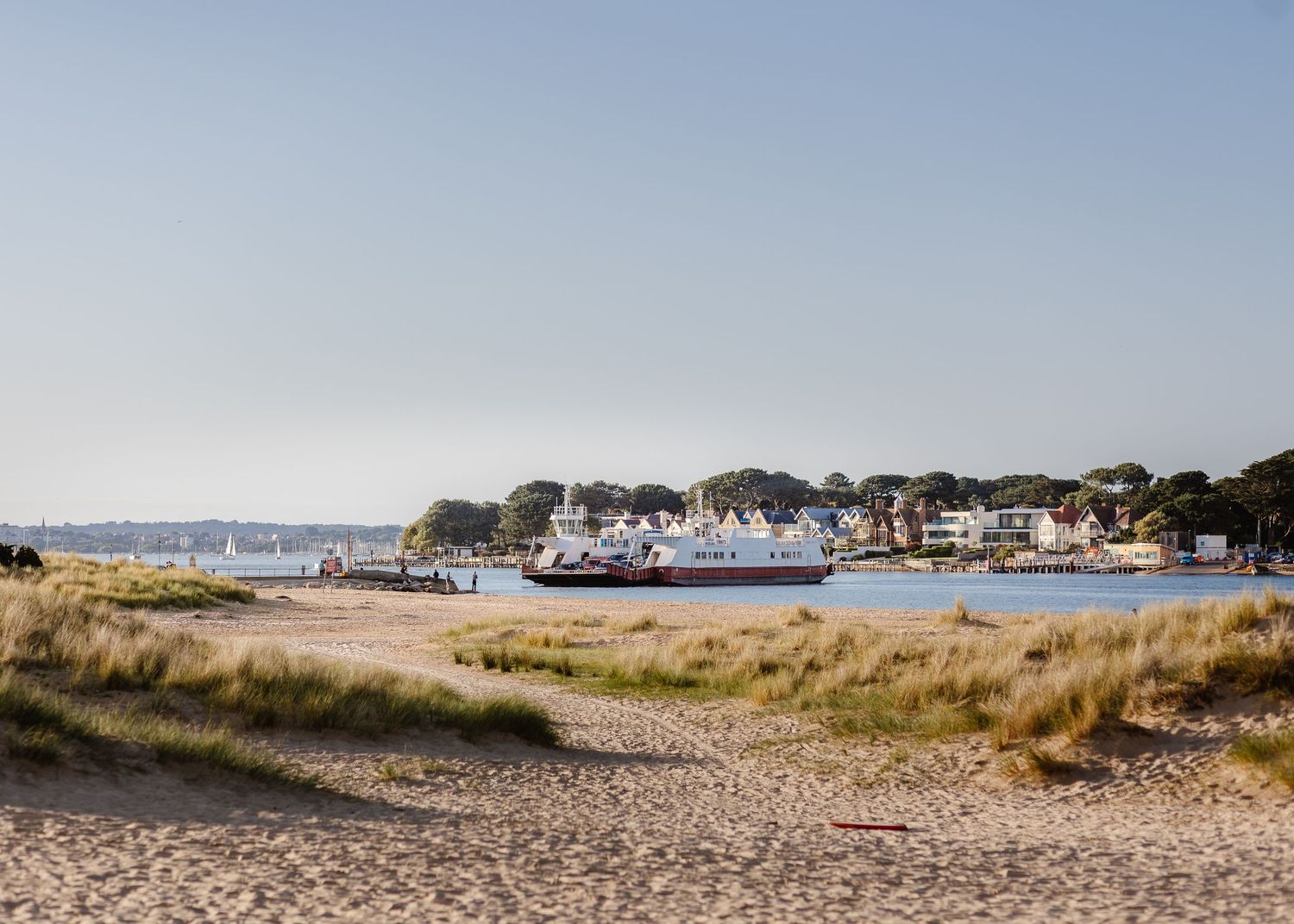 Sandy beach with dune grass leading to a calm harbor with moored boats and colorful waterfront homes under a clear blue sky.