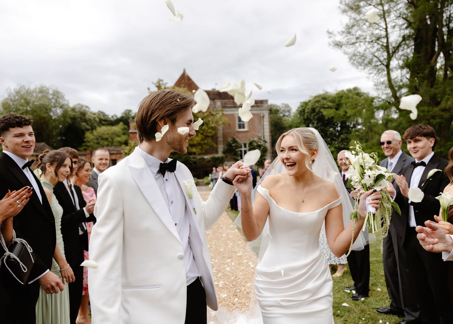 Bride and groom walk joyfully as guests toss white confetti petals outside a historic venue.
