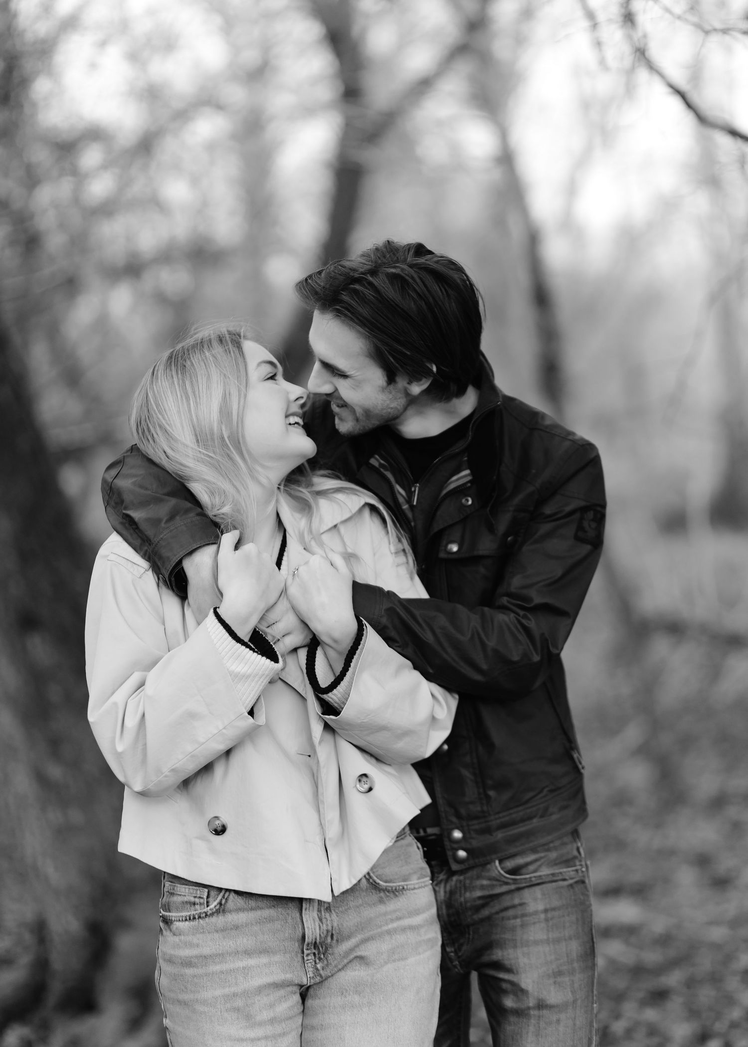 Black and white photo of a smiling couple embracing in a forest, wearing jackets, sharing a joyful romantic moment.