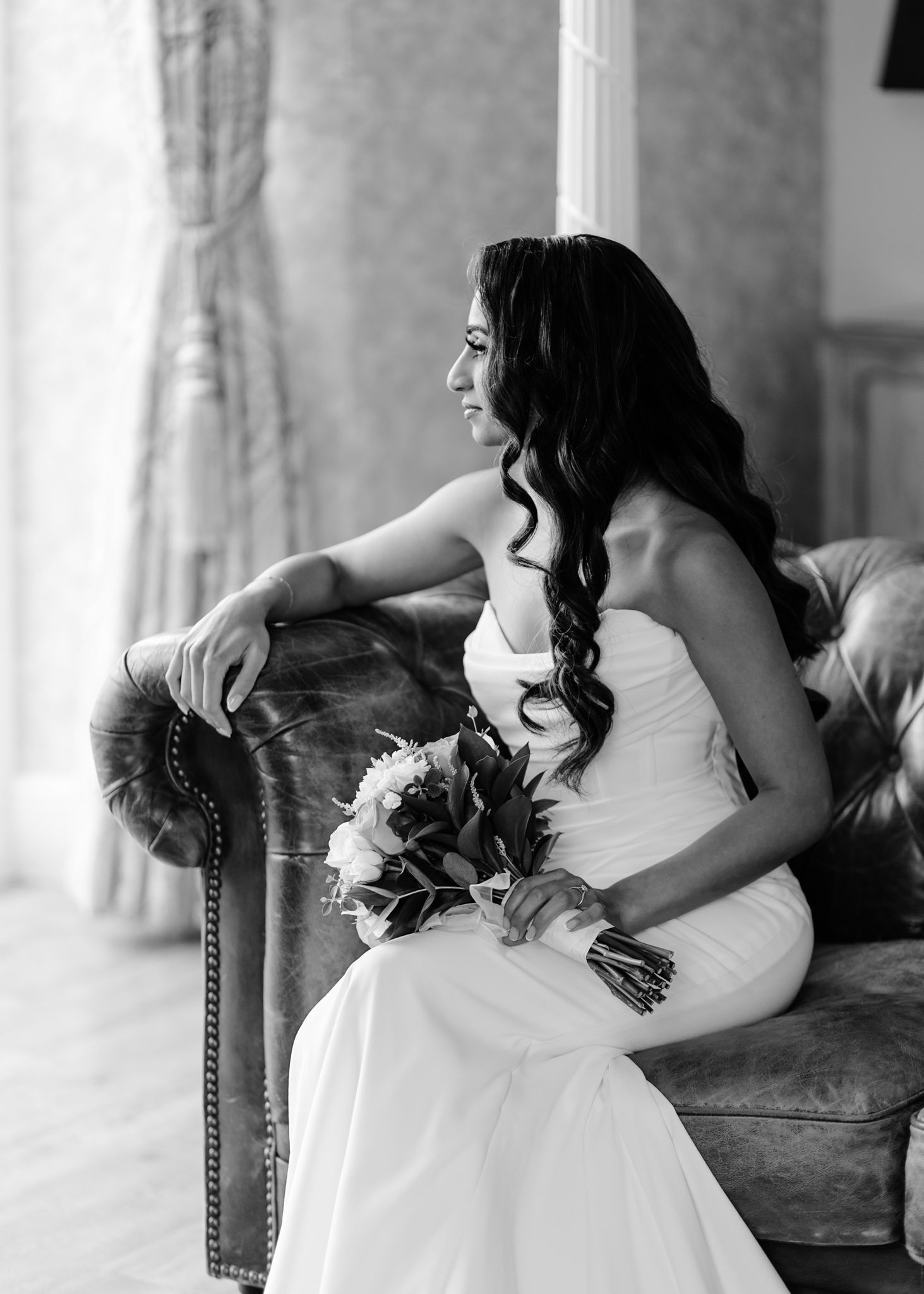 Black and white portrait of a bride in a strapless gown holding a bouquet while seated on an elegant vintage chair.