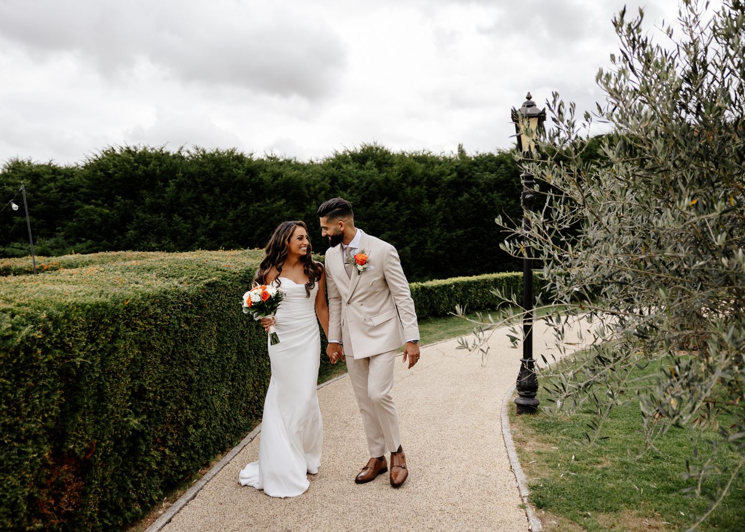 A couple walks together on a garden path surrounded by hedges on a cloudy day.