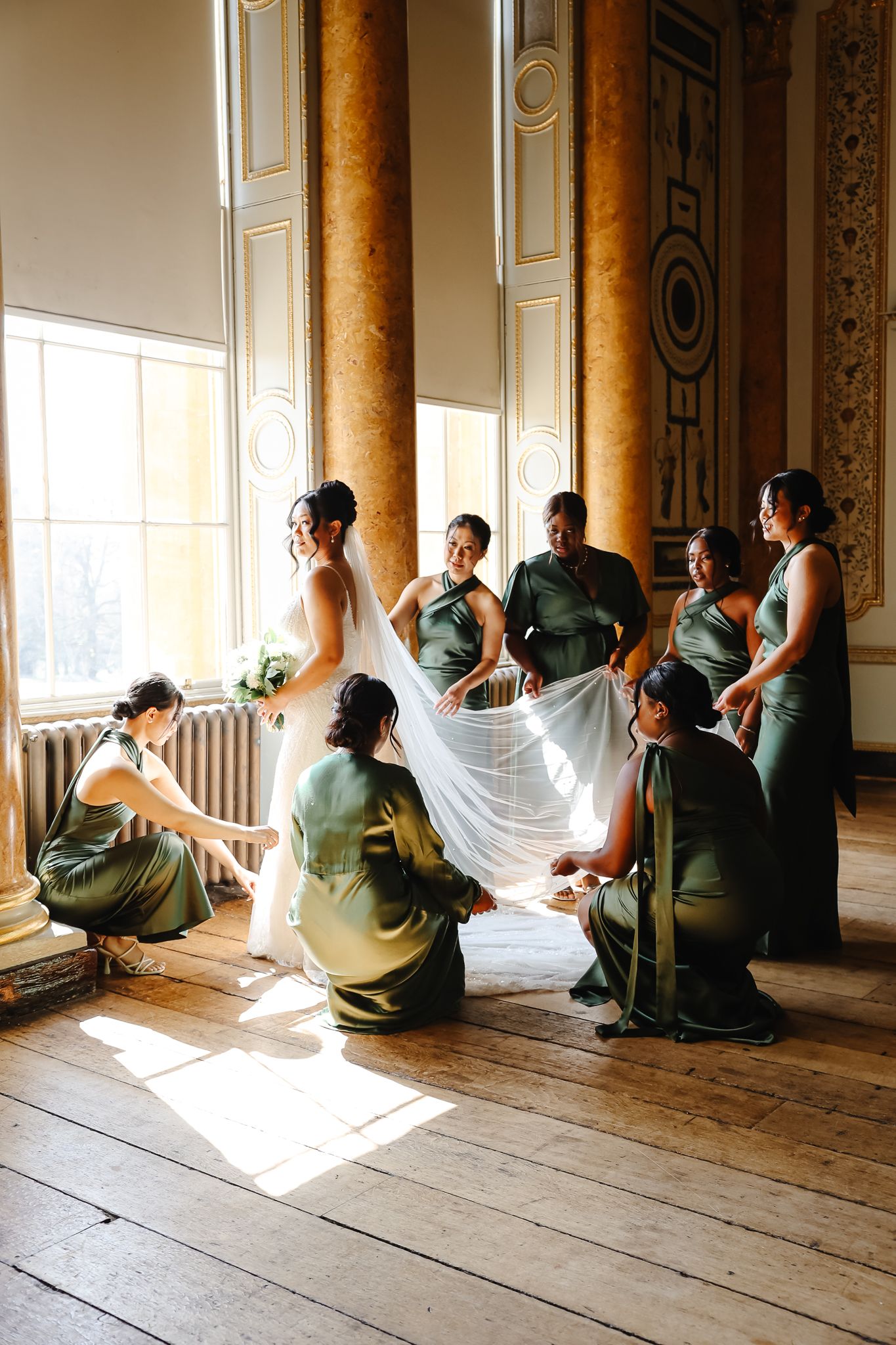 Wedding party in sage green dresses helps with white gown in elegant room with tall columns and sunlit windows.
