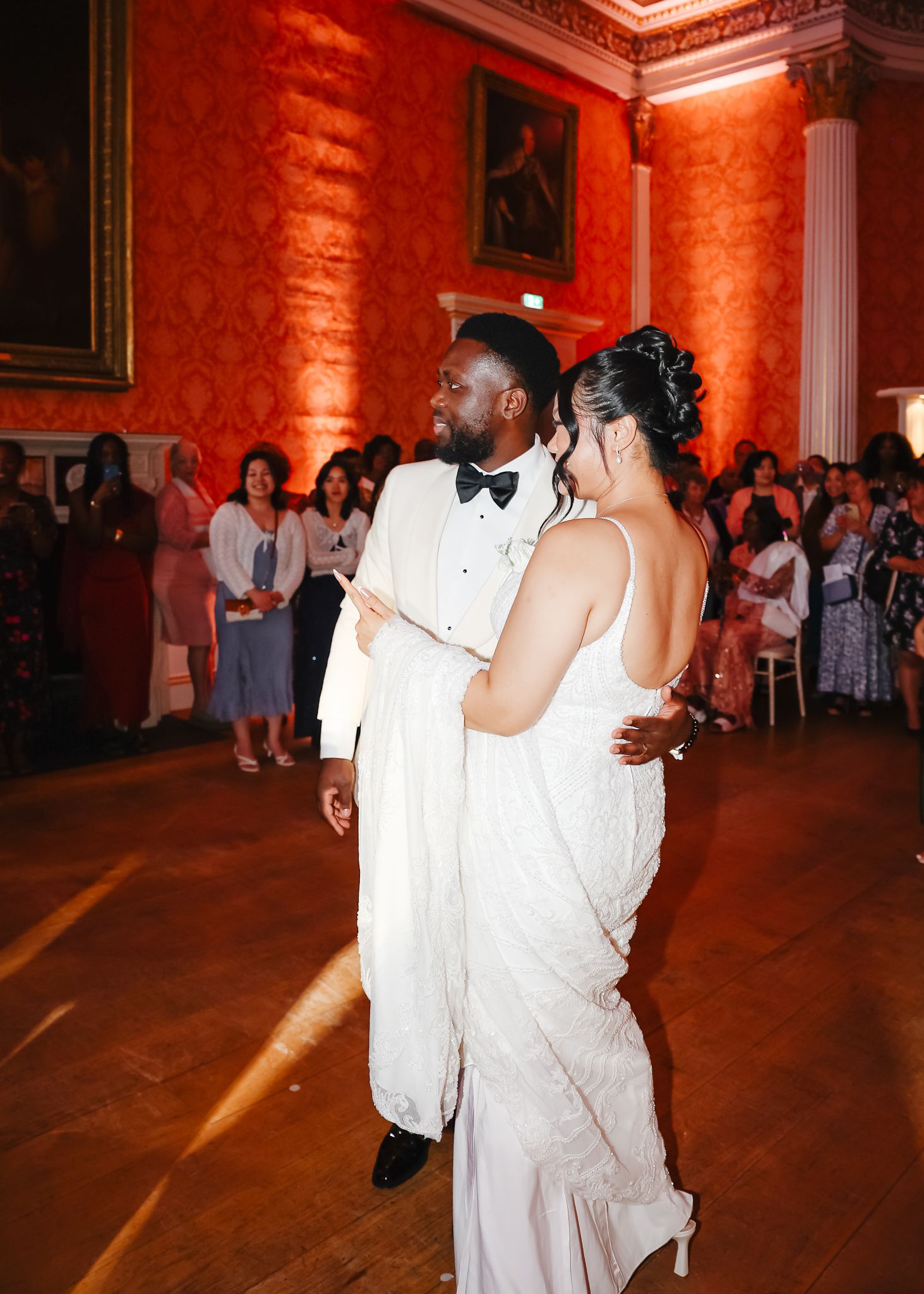 A couple in white attire share an intimate dance moment at a reception venue with warm red lighting.
